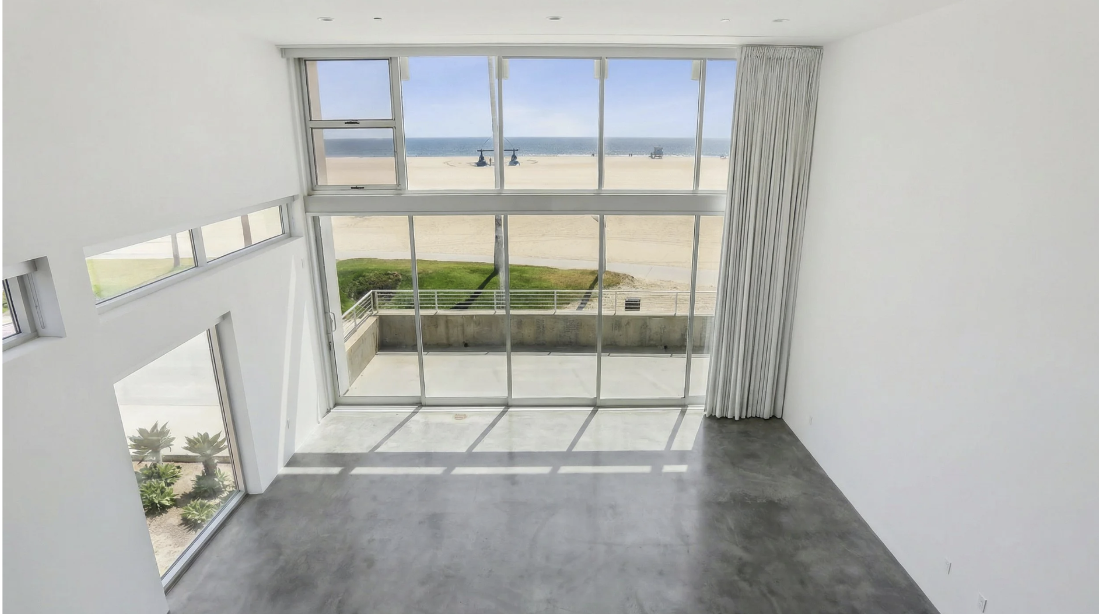Upside-down image of a modern apartment with large floor-to-ceiling windows overlooking a beach and ocean, with a balcony outside.