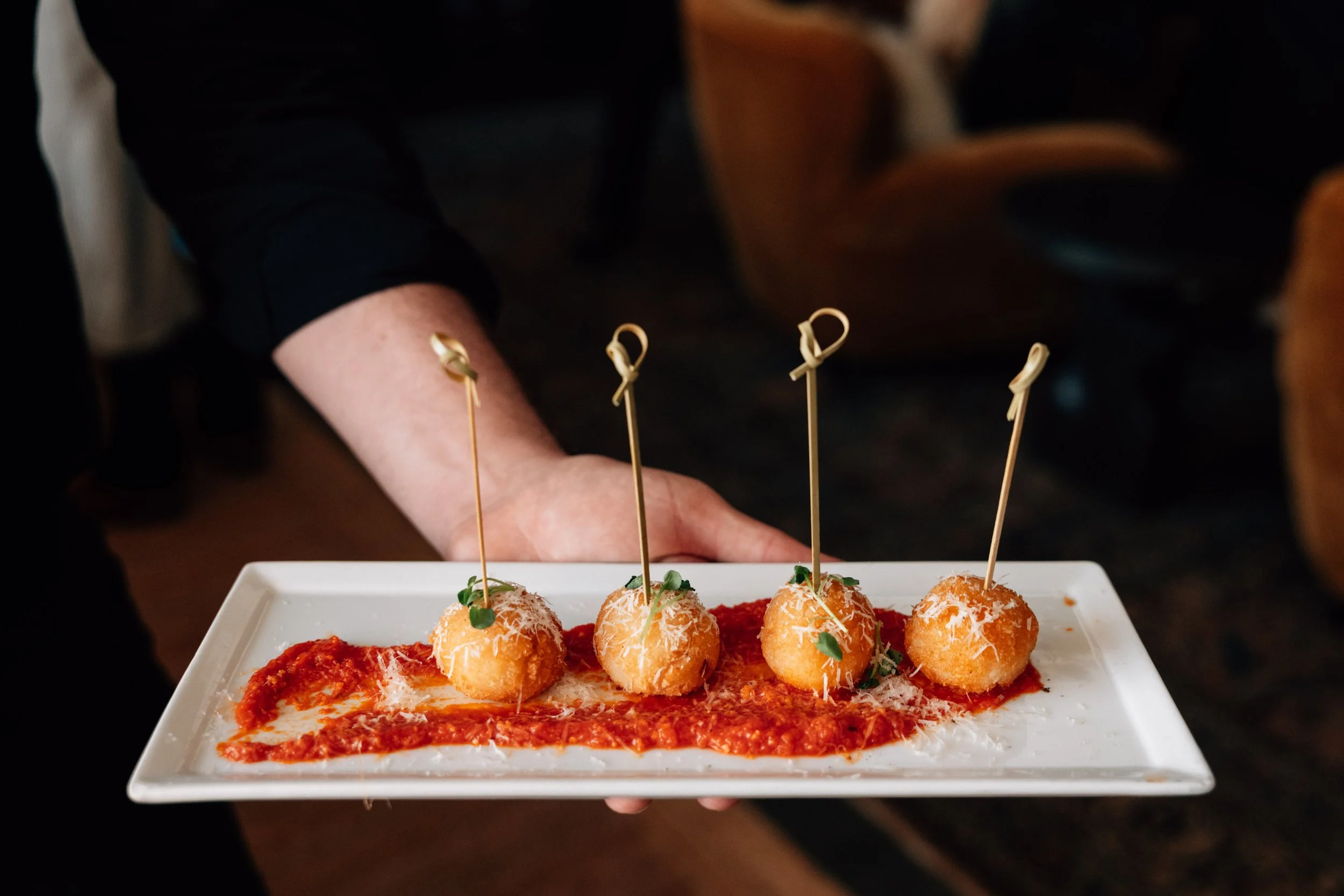 A person holding a white rectangular plate with four fried food items topped with cheese and small green garnishes, with a red sauce or paste underneath on the plate.