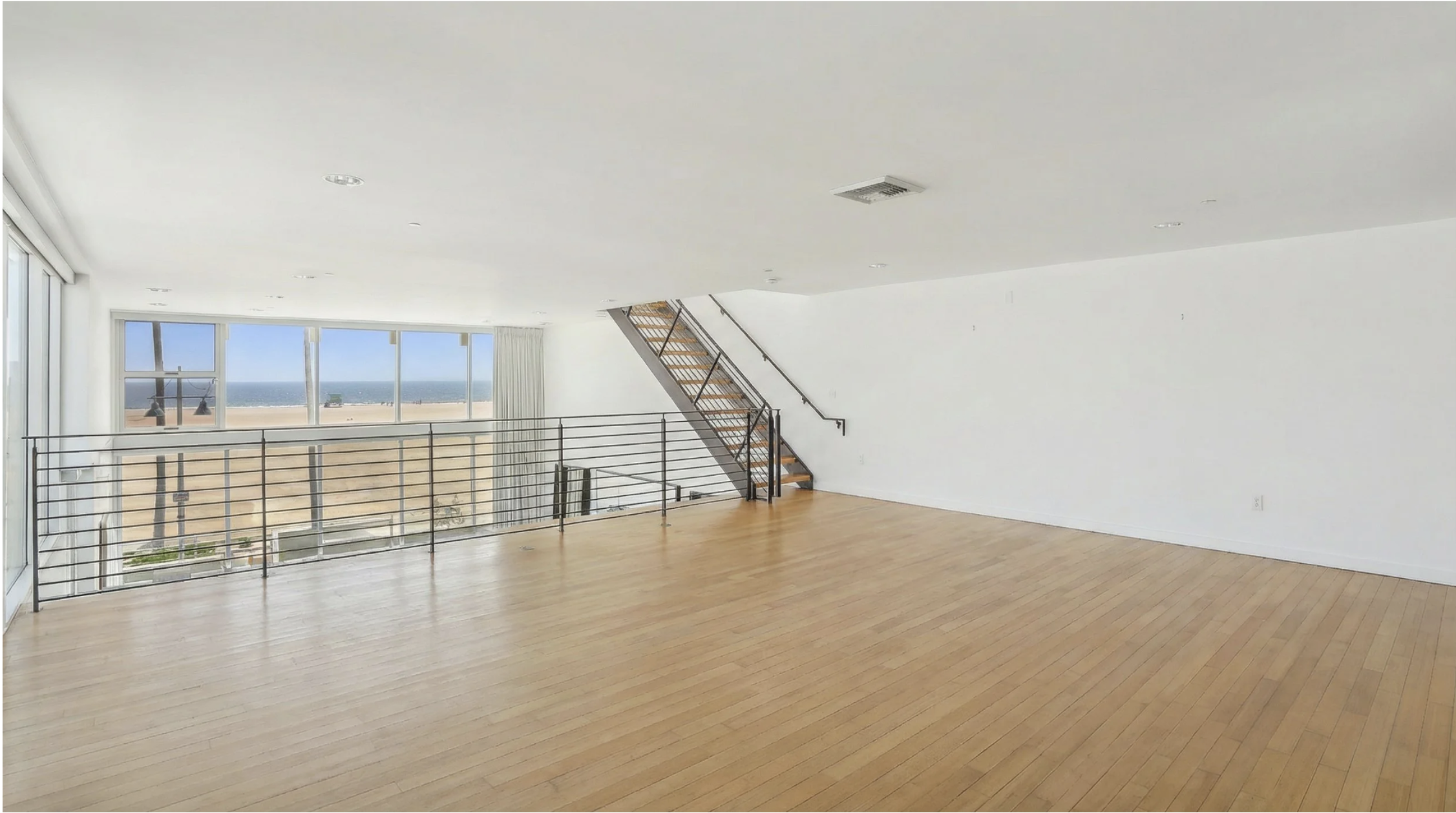 Empty modern living space with large windows showing a view of the ocean, a staircase with wooden steps and metal railing, and a light-colored wooden floor.