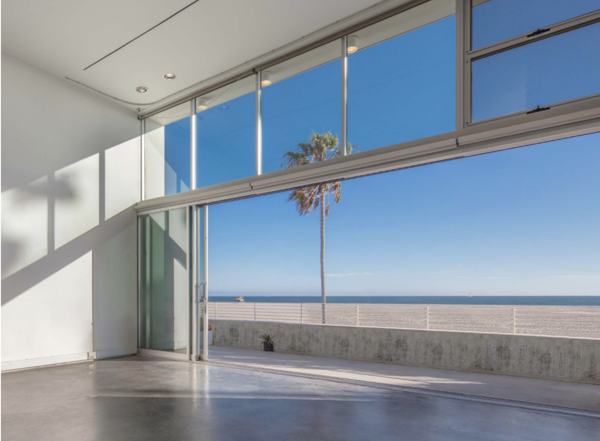 Modern interior space with large glass sliding doors opening to a beach view, revealing a sandy shore, ocean, and palm trees under a clear blue sky.