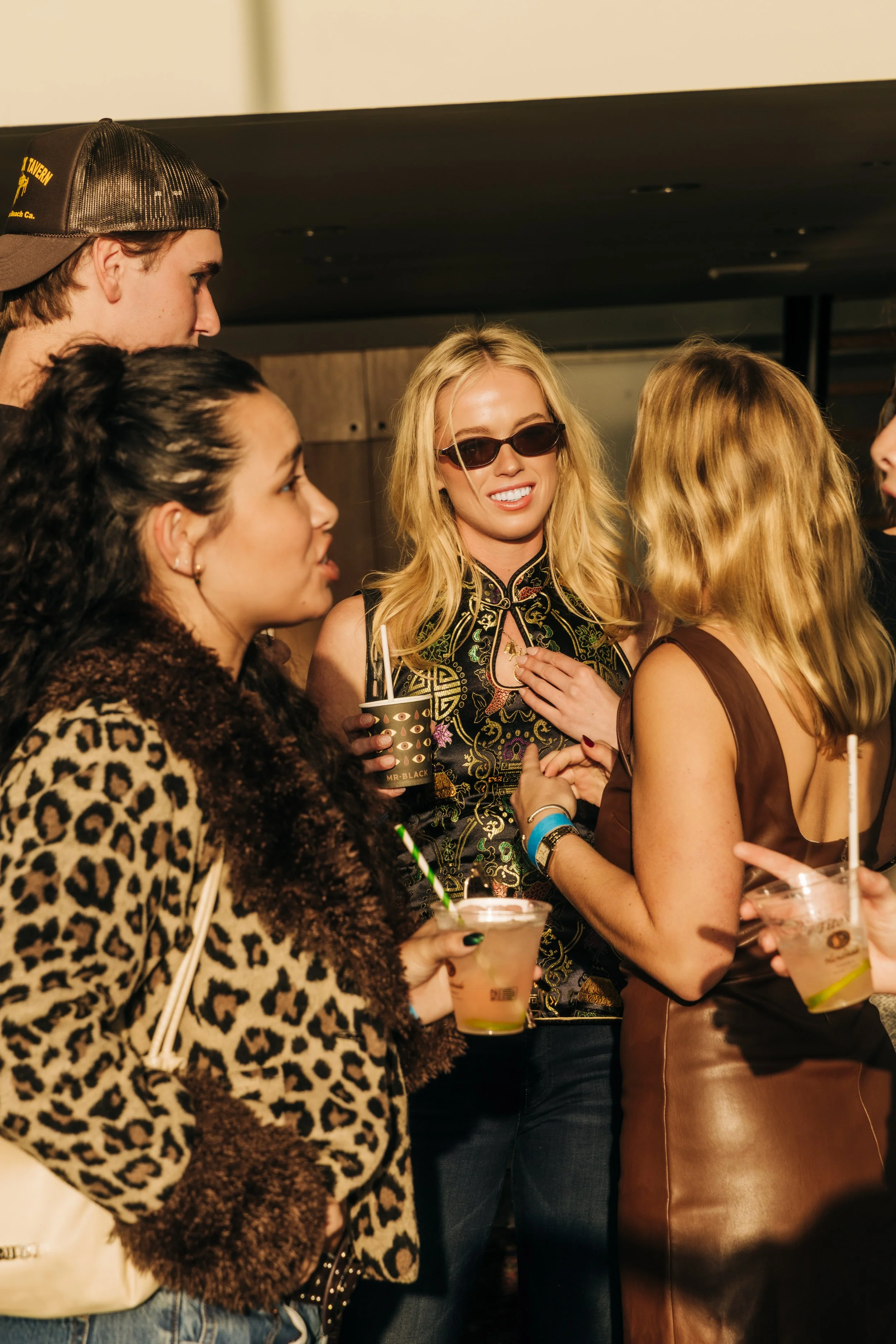 Group of people socializing, holding drinks at an indoor gathering, with women in fashionable clothing and accessories, smiling and engaging in conversation.