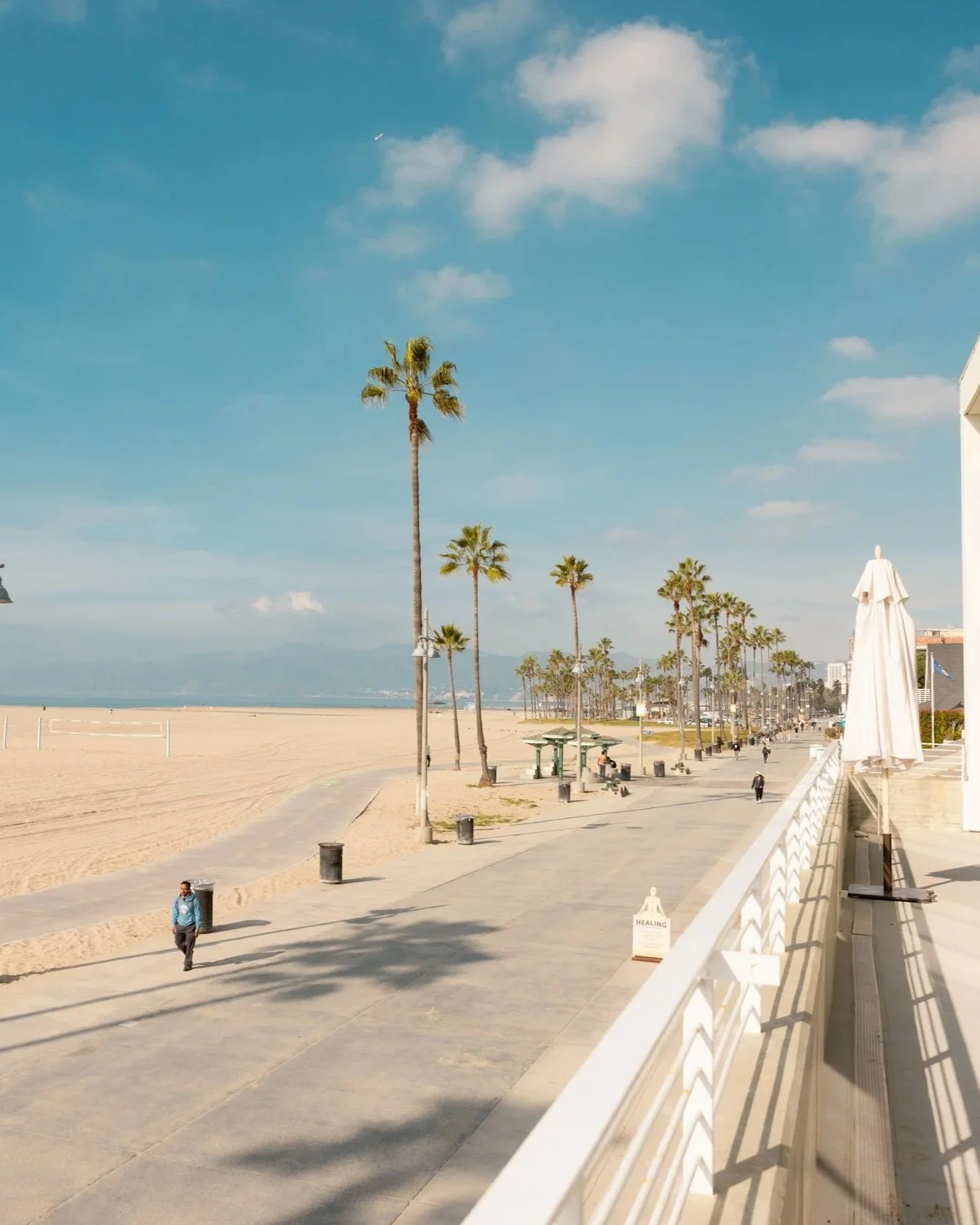 A sunny beachside boardwalk with palm trees, benches, and people walking along, with sandy beach and ocean in the background, and a clear blue sky with a few clouds.