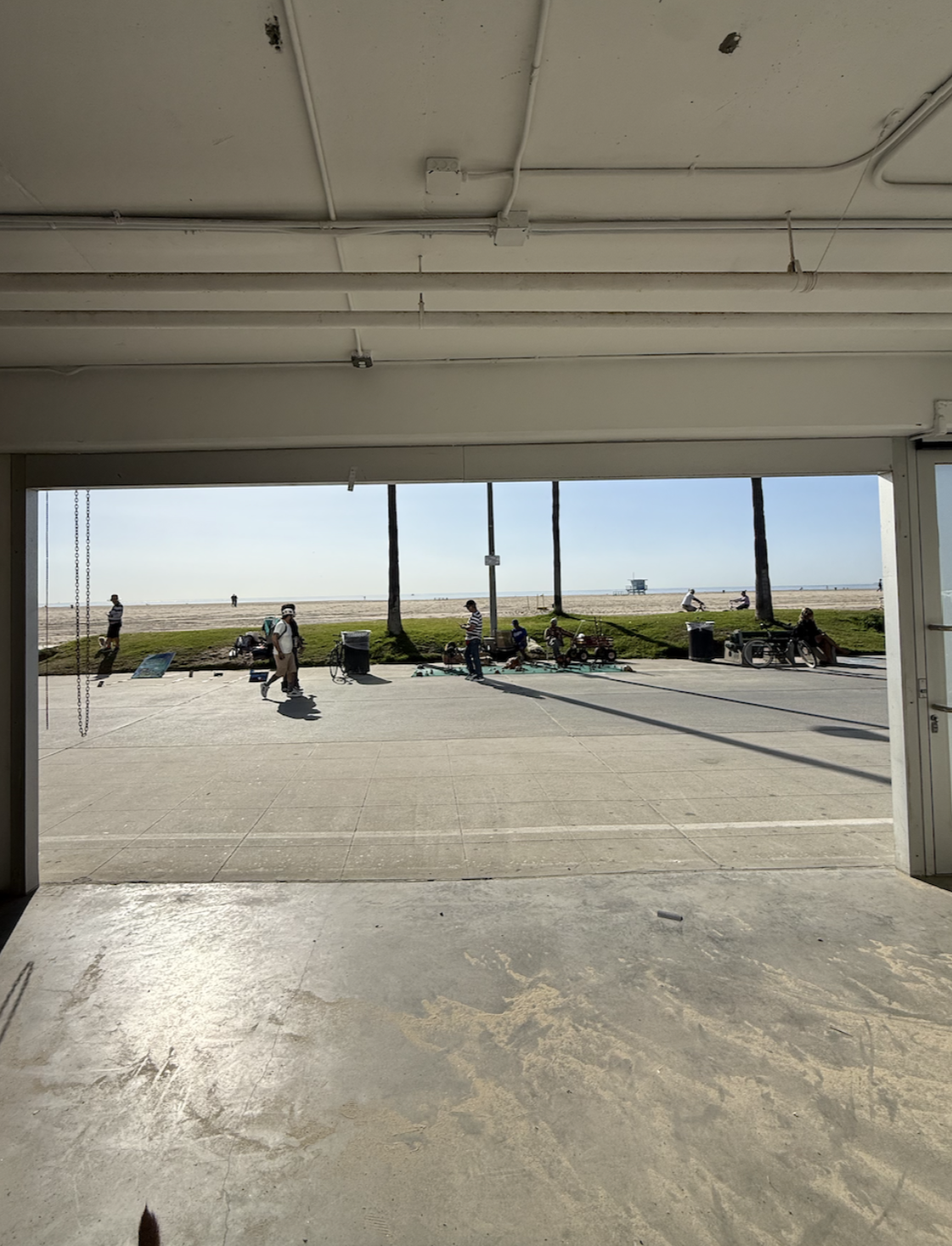 View from inside a building looking out to a beachside scene with people walking and sitting on benches, palm trees, and a lifeguard tower in the distance on the sandy beach.