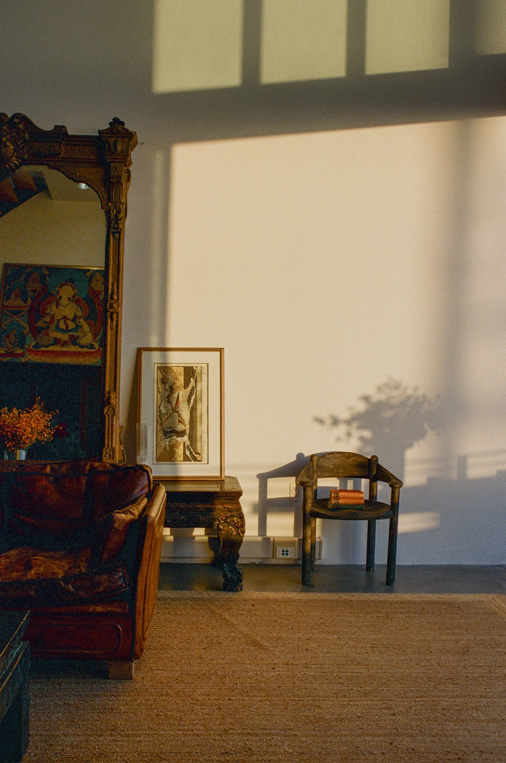 A corner of a room with eclectic furniture, including a large vintage mirror, a leather armchair, framed artwork on the wall, and sunlight casting shadows through a window.