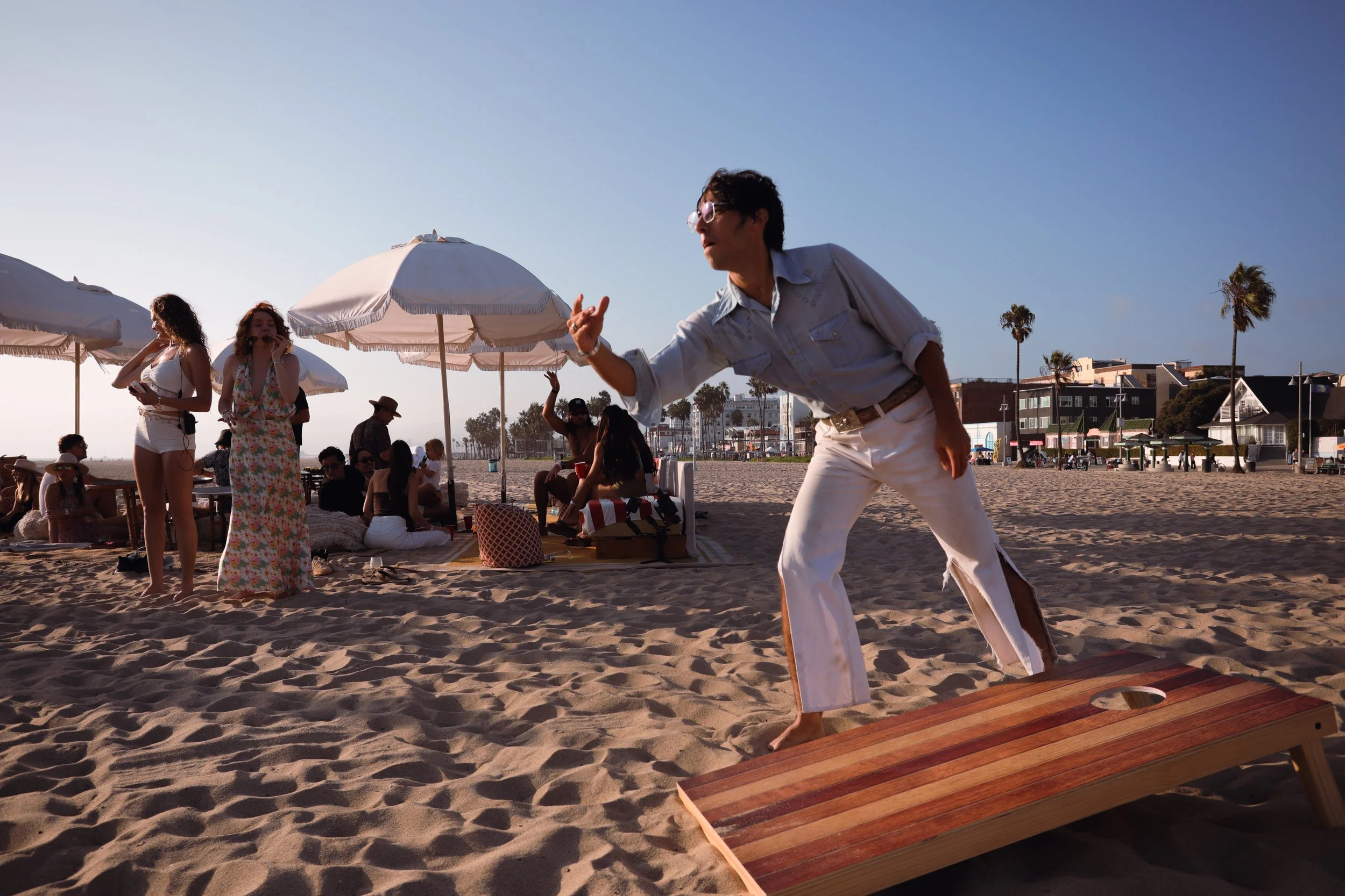 A man dressed in white pants and a light-colored shirt is performing on a wooden platform at the beach. There are white umbrellas and people relaxing and socializing on the sand, some sitting on mats or chairs, with palm trees and buildings visible i