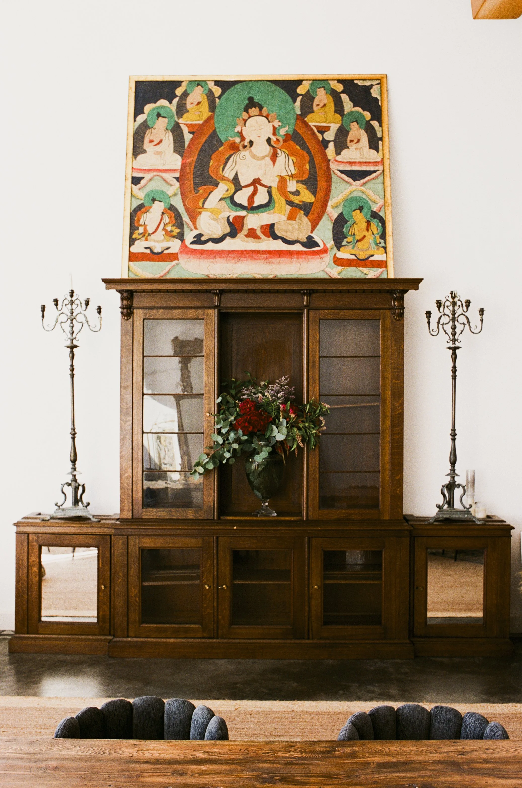 Wooden cabinet with glass doors and a vase with red and green flowers. On top of the cabinet is a pair of antique candelabras with candles, flanking a large colorful art piece of a seated deity and smaller figures, against a white wall. Dark chairs a