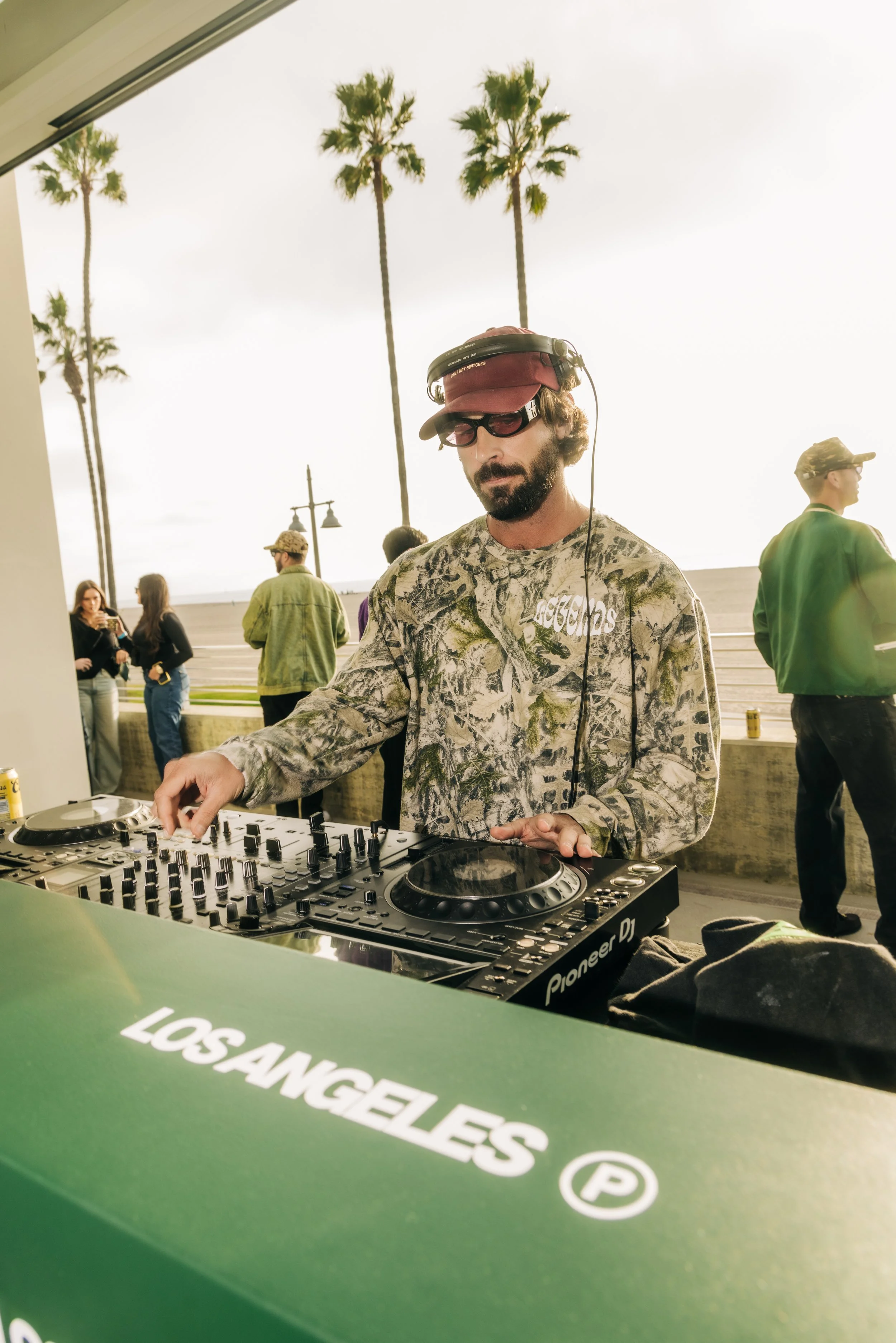 DJ performing outdoors with palm trees in the background, wearing sunglasses, a patterned long sleeve shirt, a hat, and headphones, using DJ equipment.