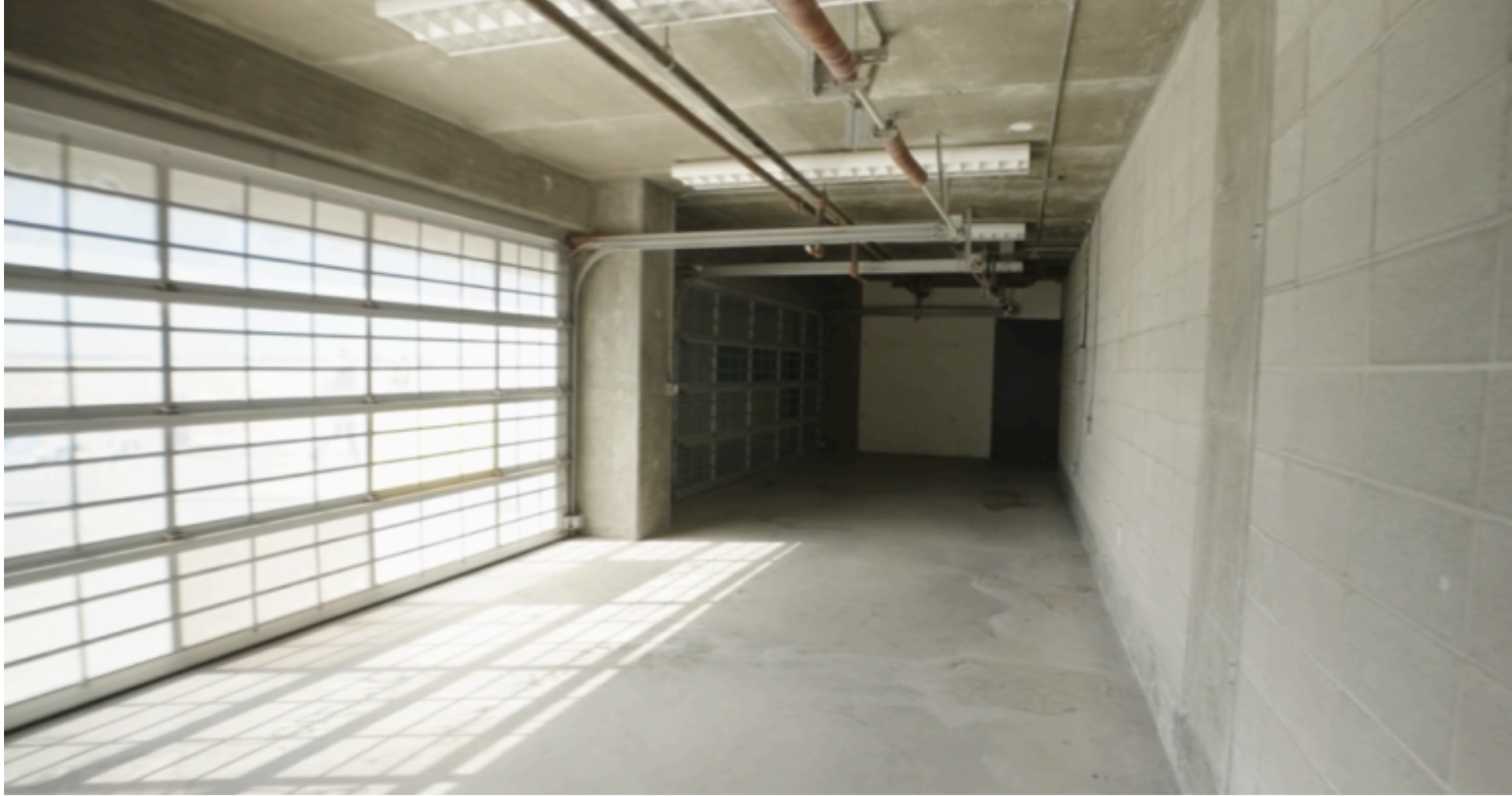 An empty, unfinished garage with a large windowed garage door on the left, concrete walls and floor, exposed ceiling with pipes and fluorescent lighting, and metal shelves at the back.