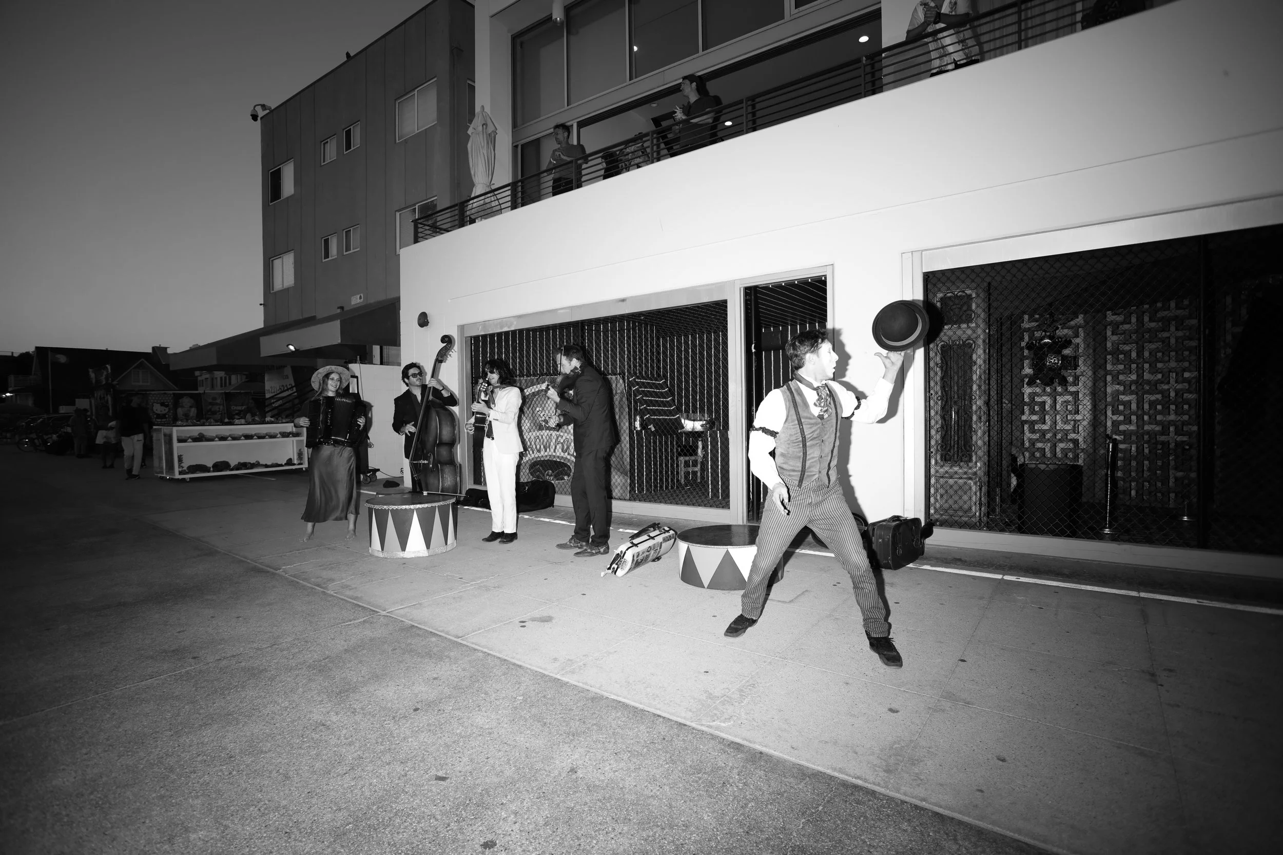 A street scene at dusk with a live outdoor band performing, including musicians playing a guitar, double bass, and accordion. A man in party attire is holding a hat in the foreground. People are on a balcony watching the performance.