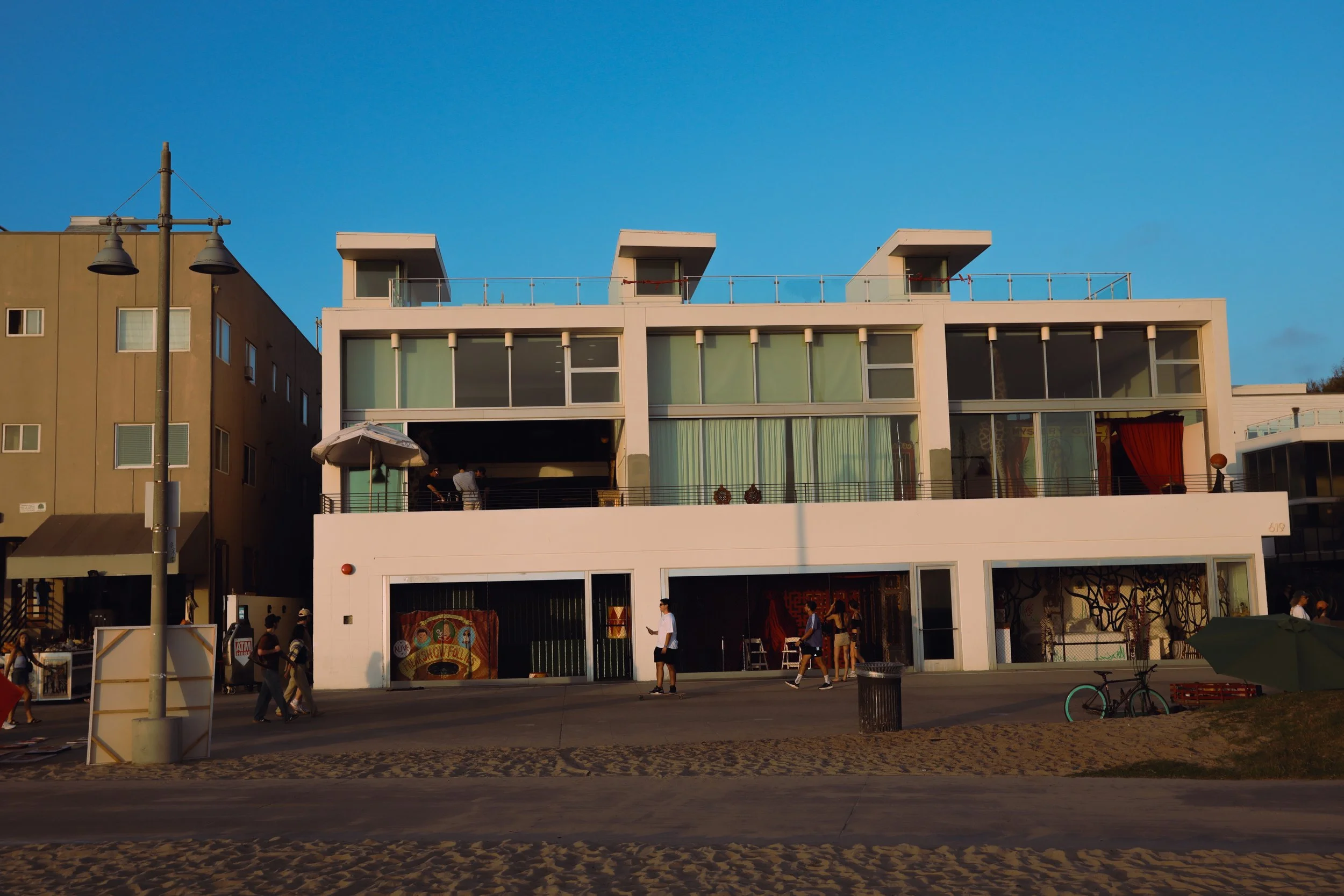 A modern multi-story building with large glass windows and a rooftop terrace, located alongside a busy street with pedestrians and bicycles in the foreground, under a clear blue sky.