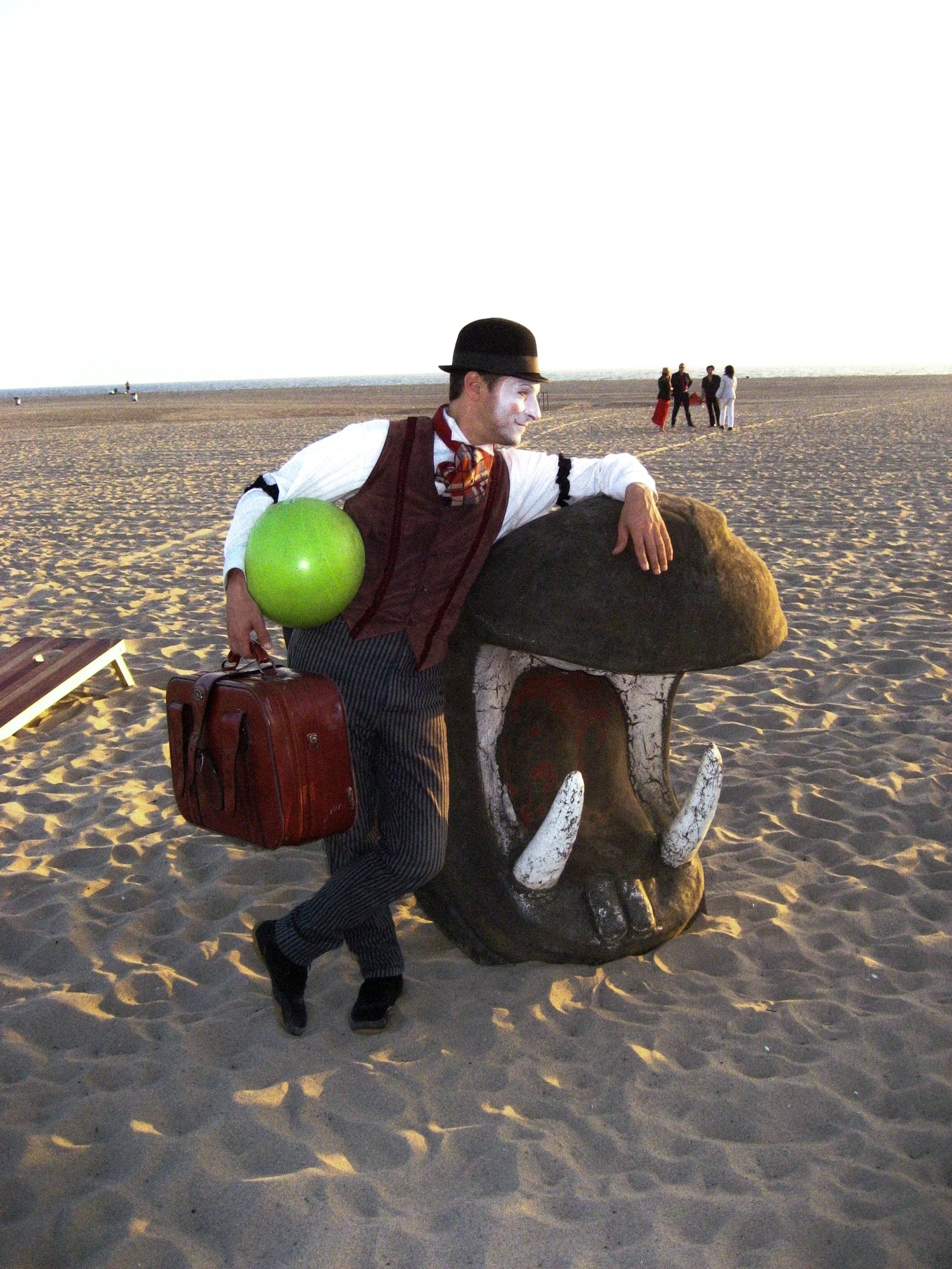 A mime dressed in old-fashioned clothing, holding a green ball and a suitcase, leaning on a large animal skull on a sandy beach during sunset, with a small group of people in the background.
