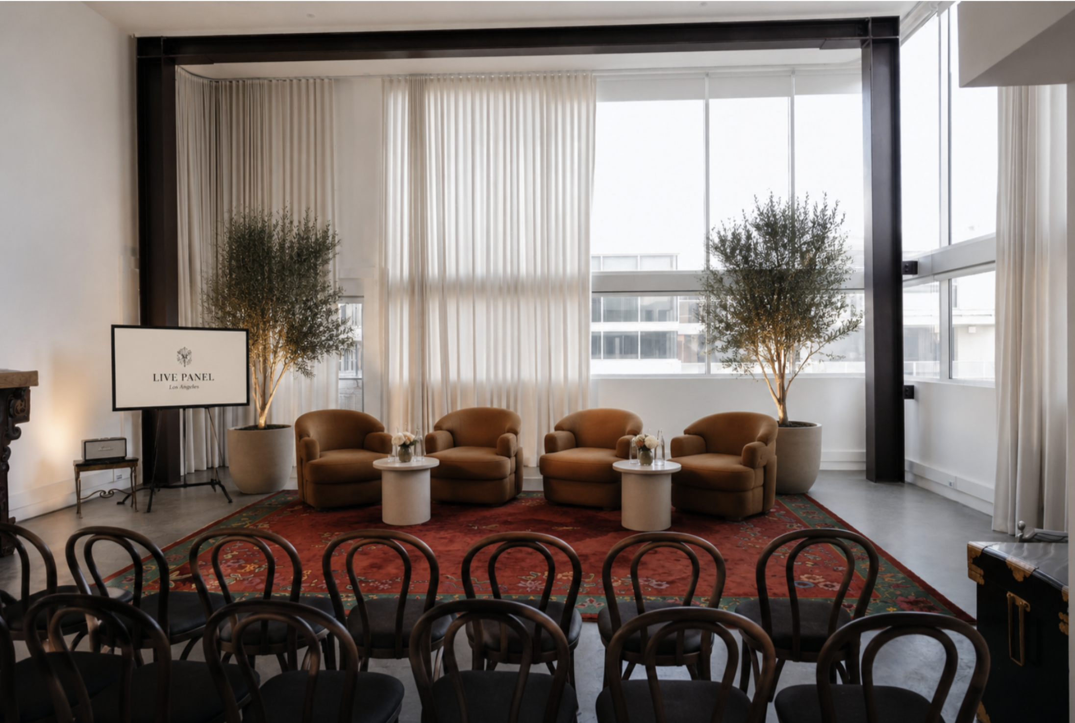 Indoor event space with a row of black chairs at the front and a seating area with four beige armchairs, three round white tables, and potted plants. Large windows with sheer white curtains allow natural light, and a sign reads 'Live Panel Los Angele