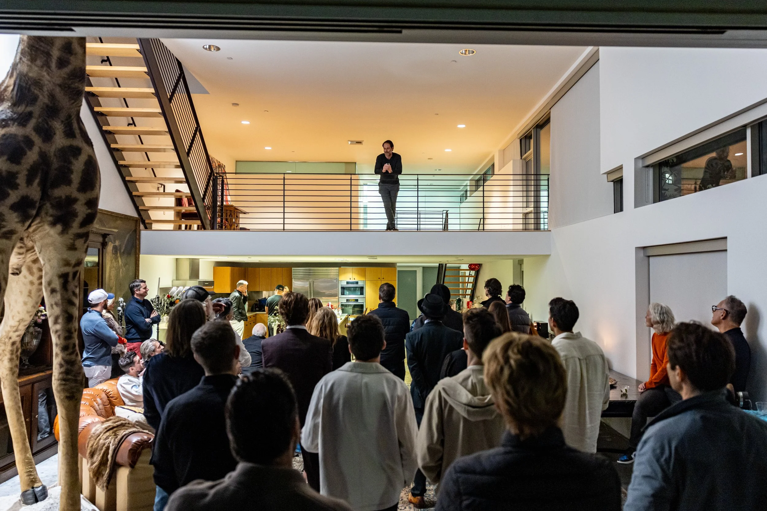 A man stands on a second-floor balcony, looking at his phone, overlooking a crowd gathered in a spacious, modern indoor event space with high ceilings and large windows.