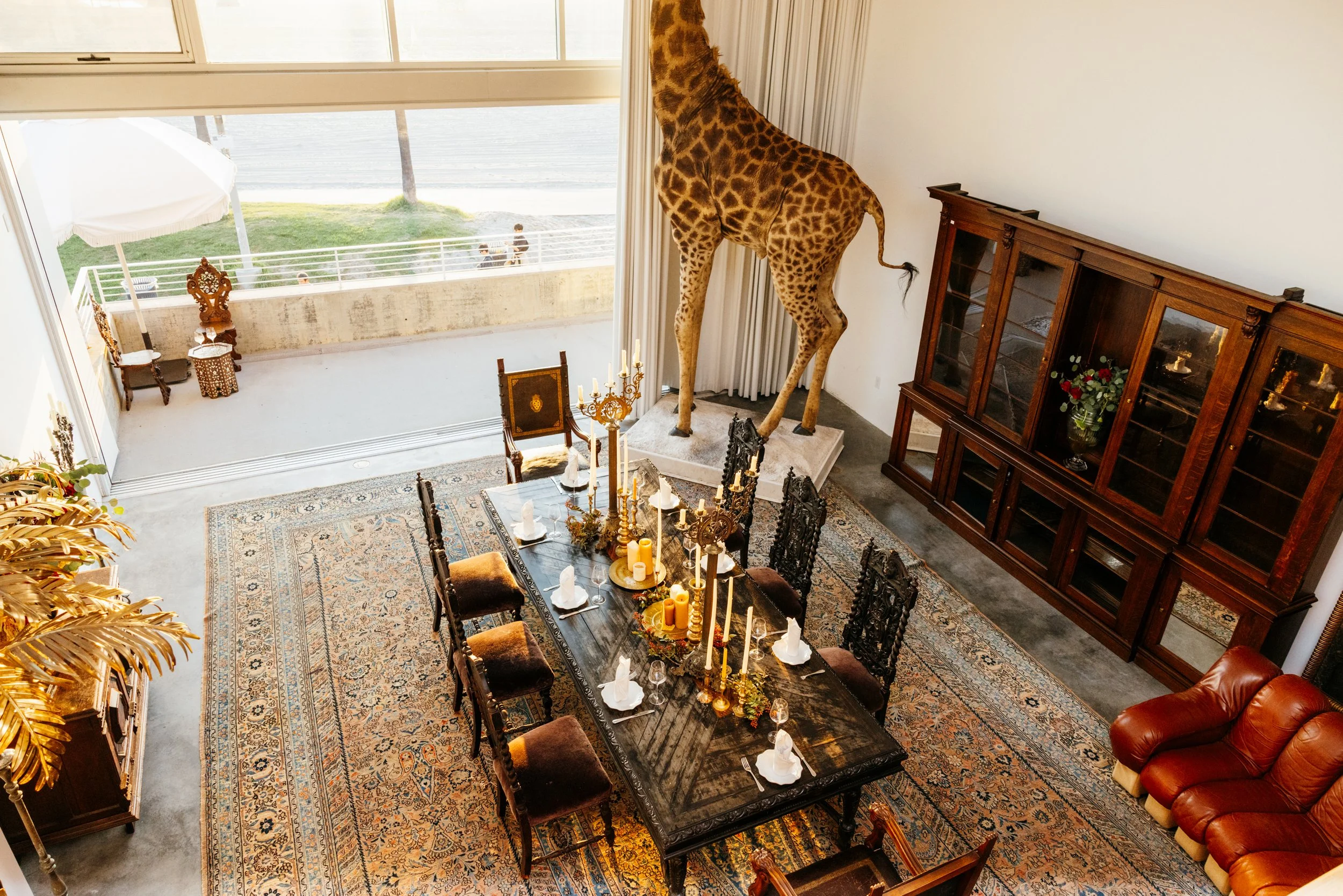 Interior view of a dining room with a large dark wood table set for a meal, including candles, wine glasses, and cloth napkins. The room features a Persian-style rug, ornate chairs, a large wooden display cabinet, and a prominent giraffe statue near the window. Outside the large windows, there's a balcony with outdoor chairs and a view of a grassy area and street with pedestrians.