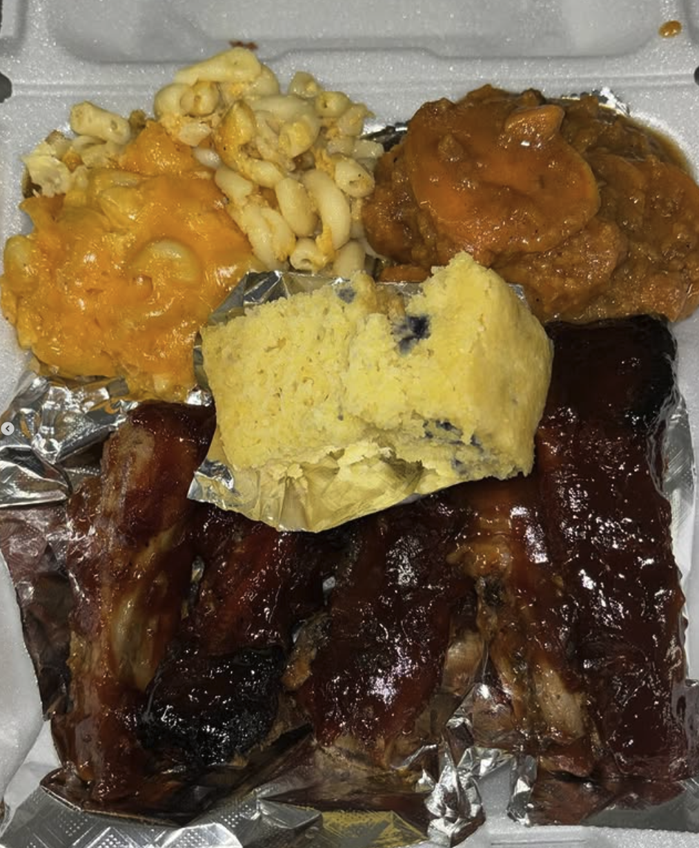 Barbecue ribs with barbecue sauce, cheddar cheese macaroni and cheese, macaroni salad, mashed potatoes, and a slice of cornbread in a foam takeout container.