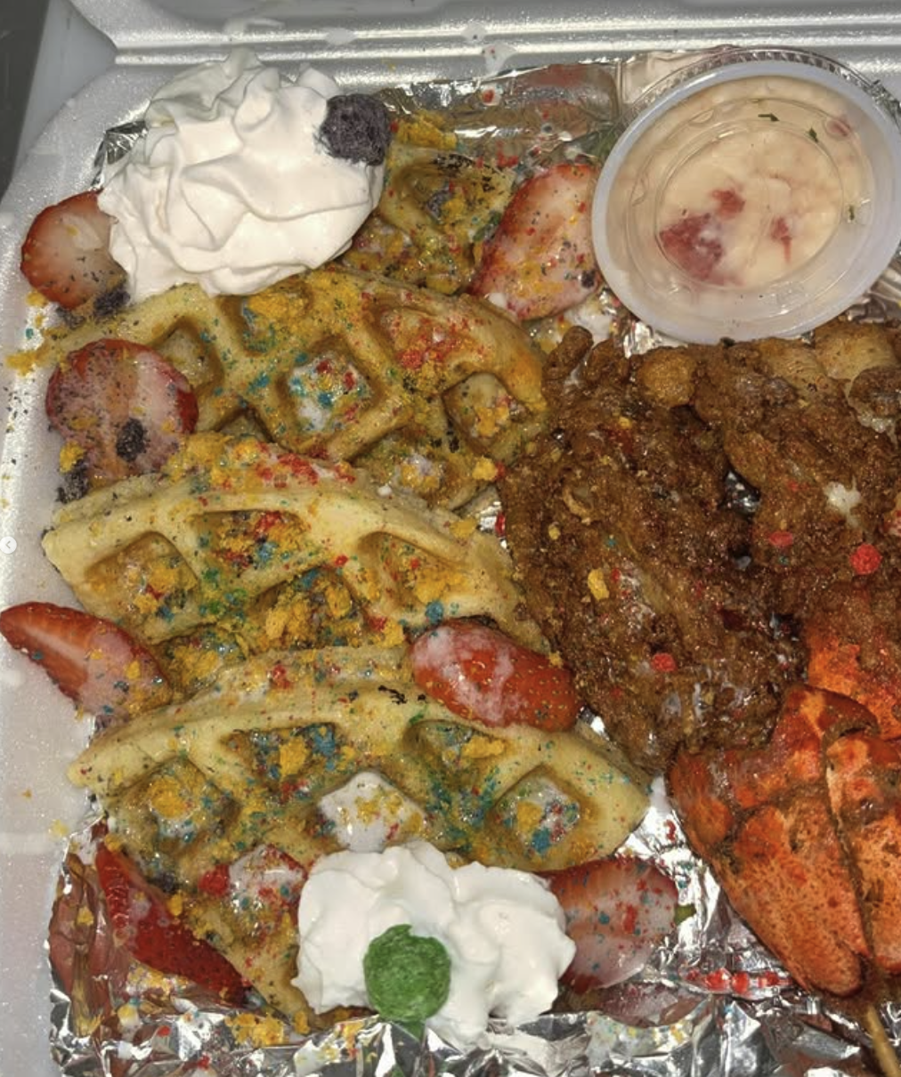 A seafood platter with fried lobster tail, cheese ravioli with colorful sprinkles, strawberries, dollops of sour cream, and a small cup of dipping sauce on foil