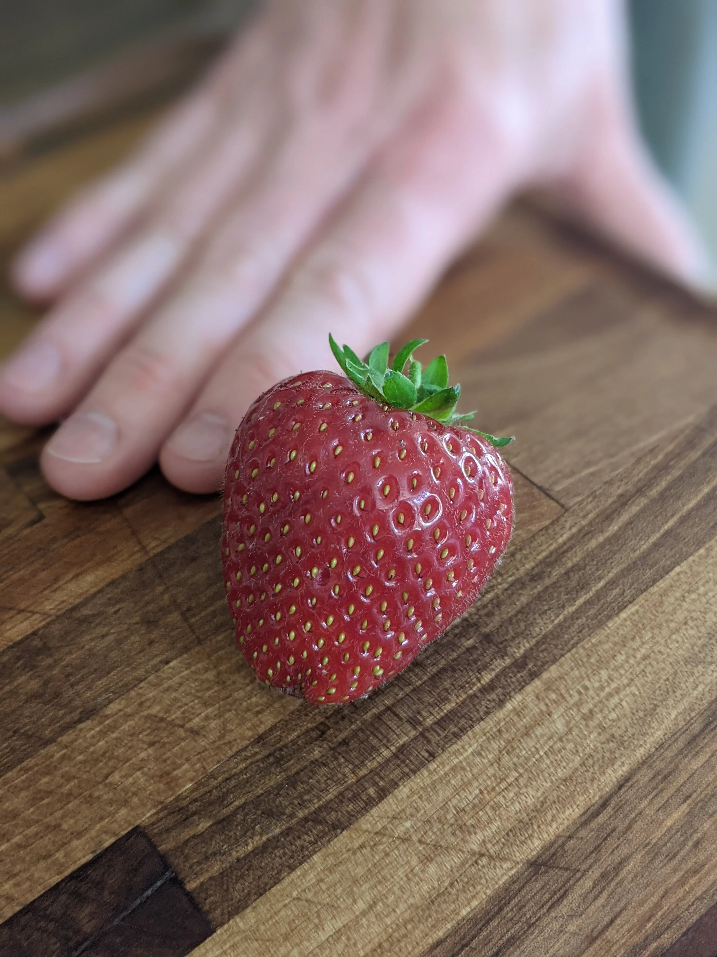 A ripe red strawberry with green leaves on a wooden cutting board, with a blurred hand in the background.