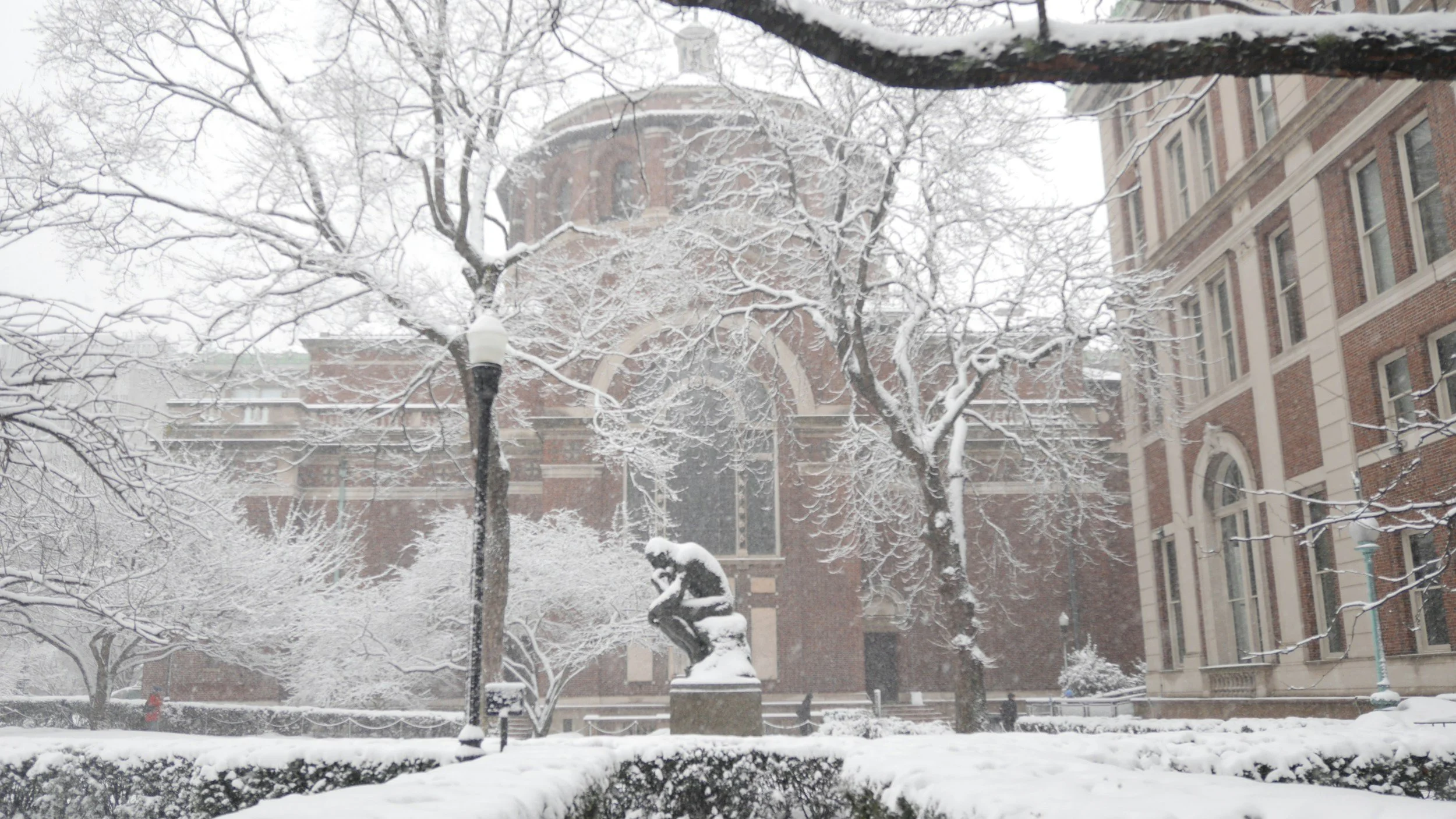Snow-covered trees, a statue, streetlamp, and brick buildings in a city scene during winter.