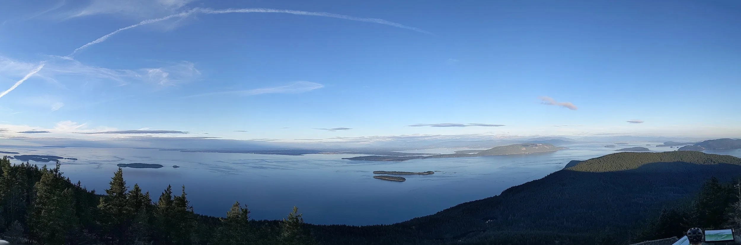 A panoramic view of the Rosario Strait, Lummi Island, and Bellingham Bay surrounded by forested hills and mountains under a clear blue sky with scattered clouds.