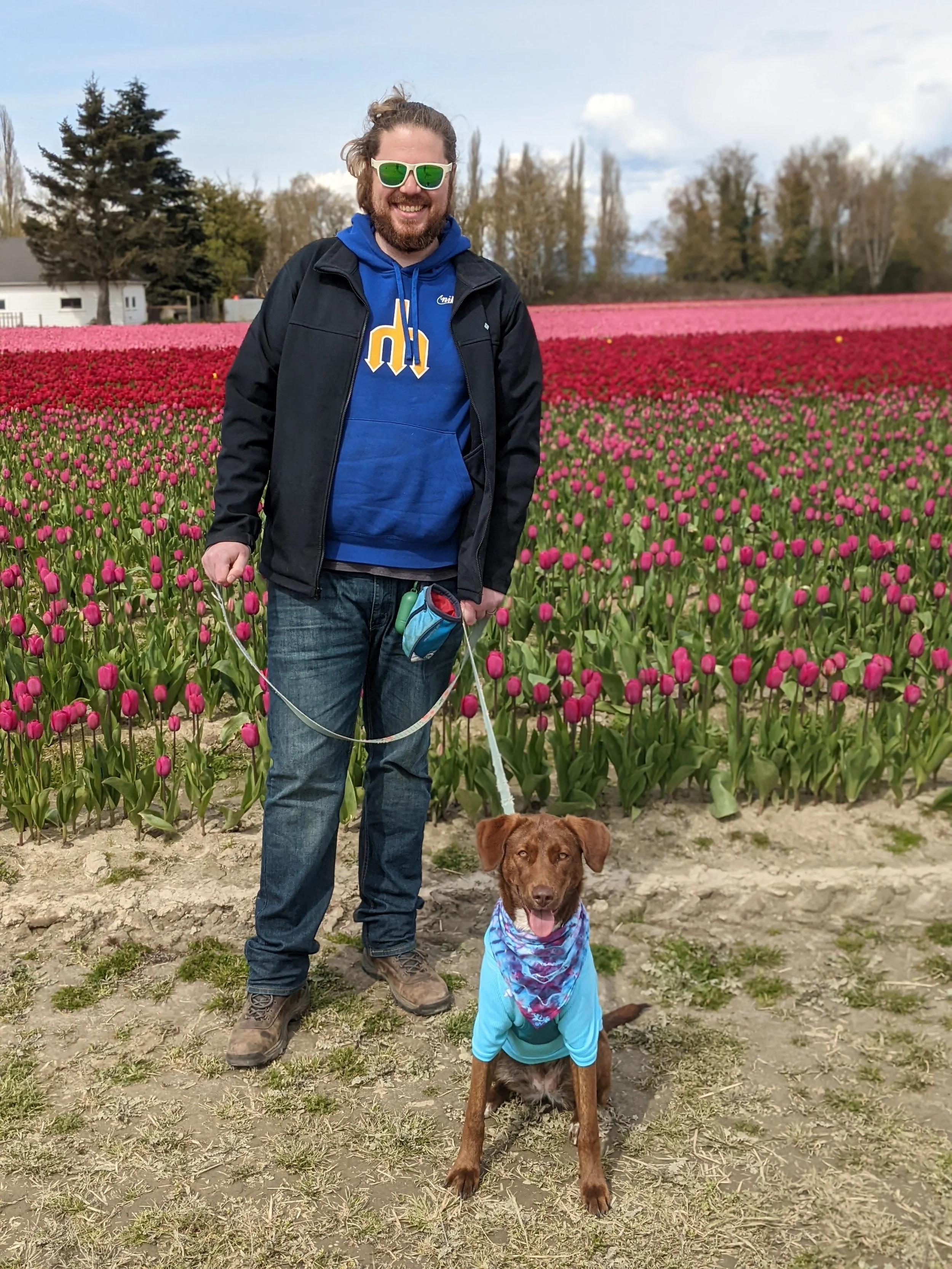Andrew Myers standing in a tulip field with a Hazel (their brown dog) sitting in front of him. Andrew is wearing a blue hoodie, a black jacket, and jeans, and their dog is wearing a blue bandana.