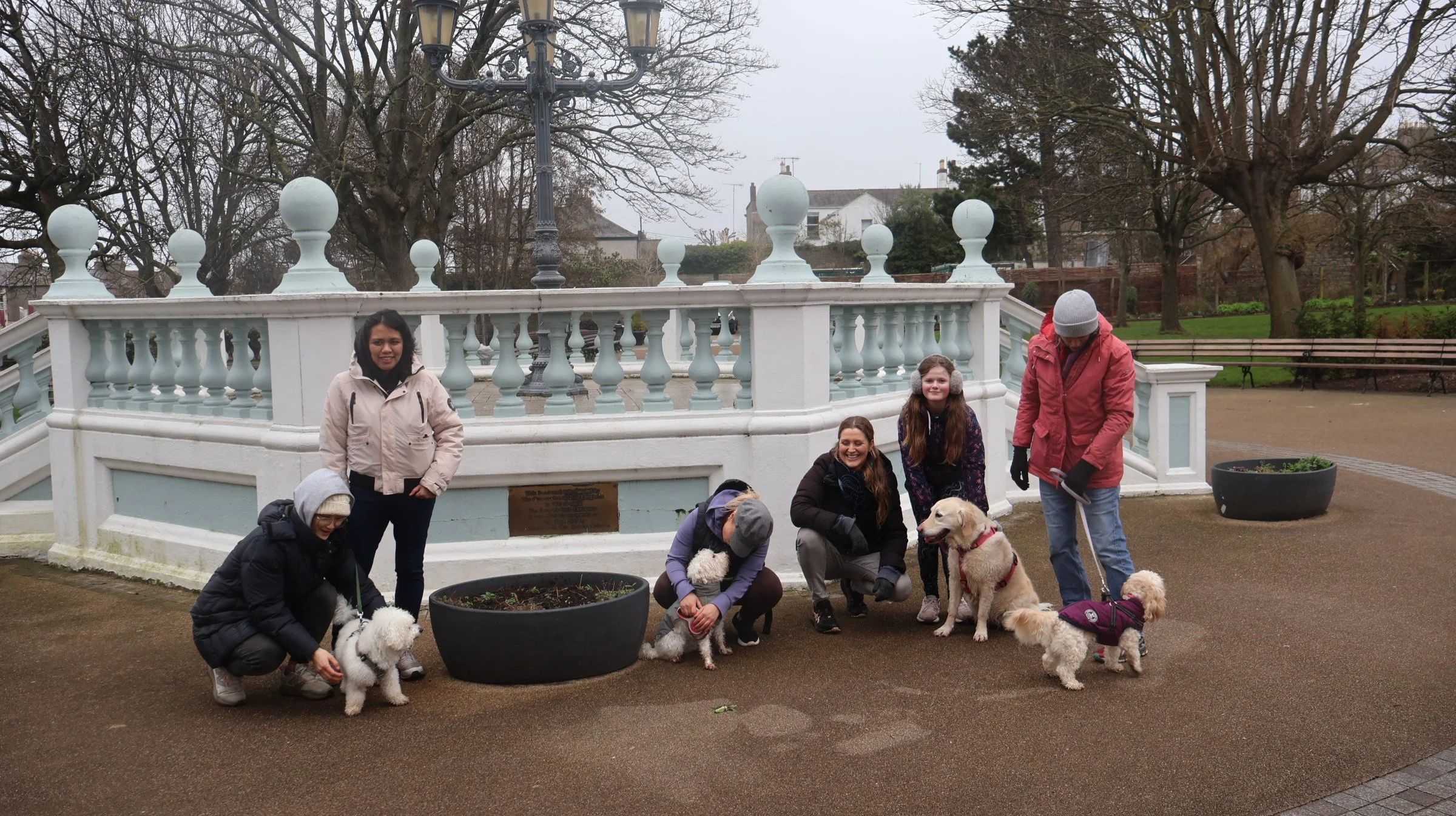 A group of five women and two children gathered outdoors in front of a white decorative bridge in a park, along with several dogs. The women are smiling and some are crouching or kneeling while interacting with the dogs. The scene appears to be on a 