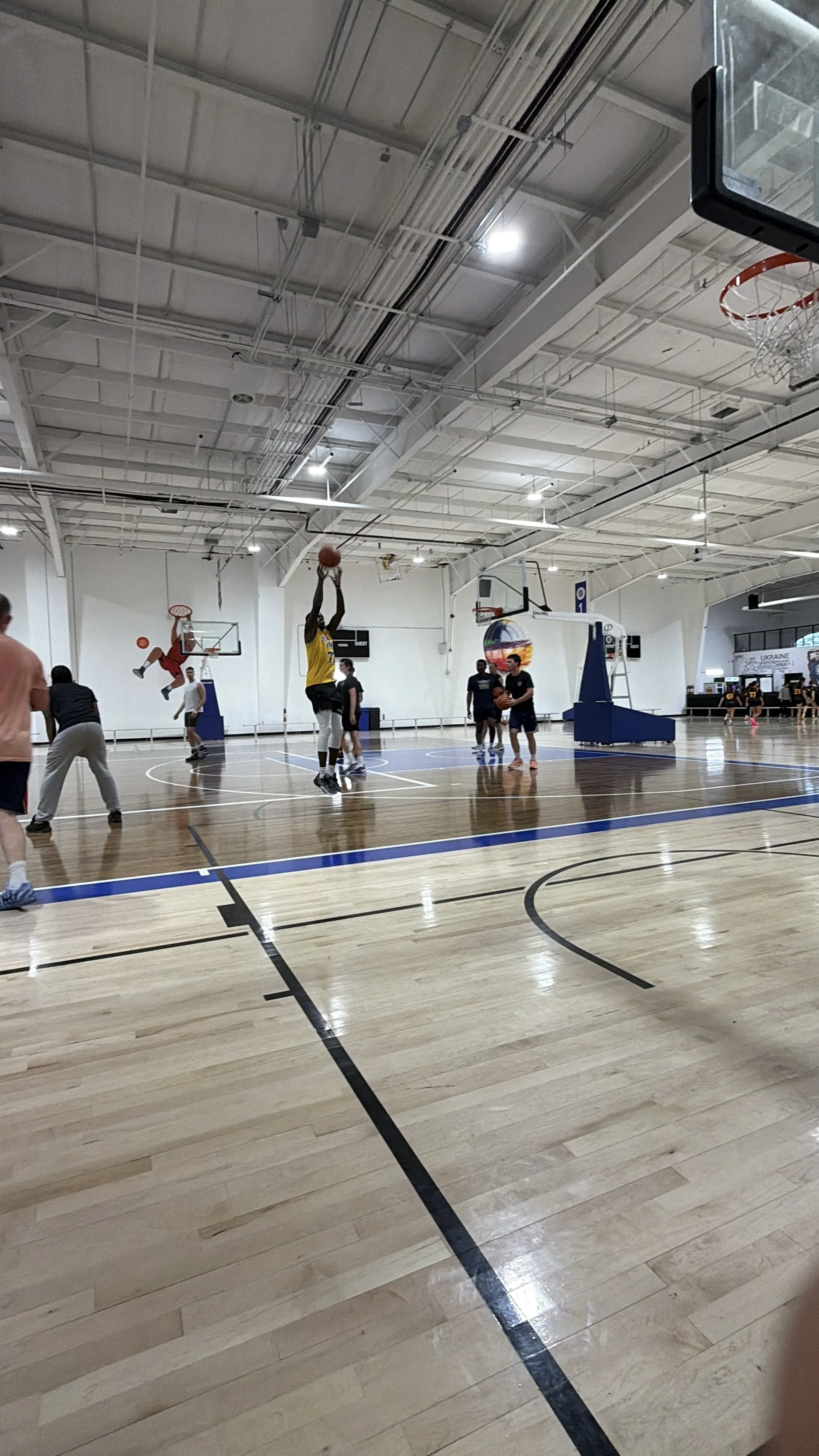People playing basketball in an indoor gym, with some players shooting and others watching.