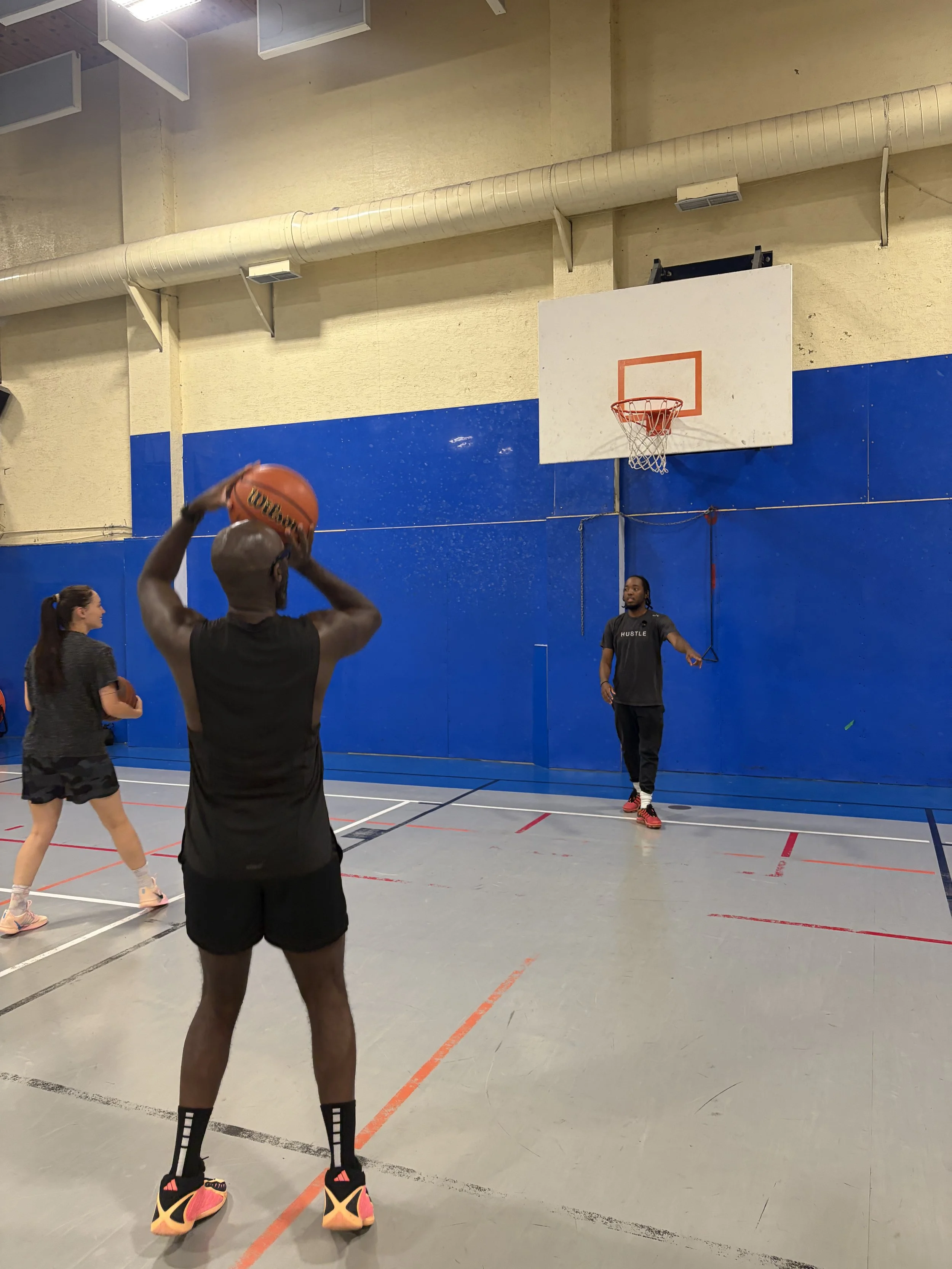 A basketball game in progress at an indoor gym. A player is preparing to shoot, surrounded by other players, with a coach on the side. The gym has a blue wall, a basketball hoop, and a ventilation duct overhead.
