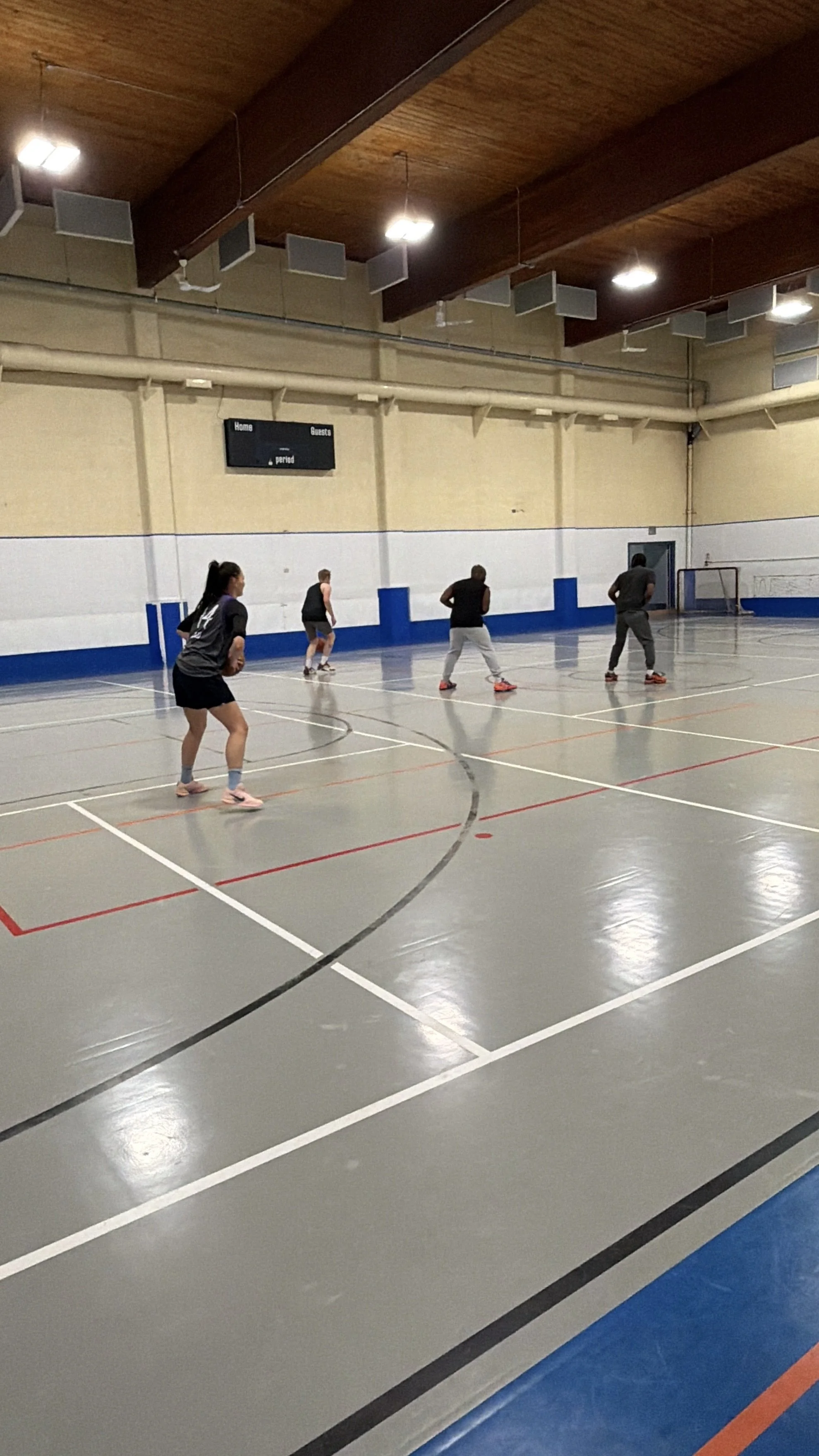 People playing basketball on an indoor court with white, blue, and orange lines and a scoreboard in the background.