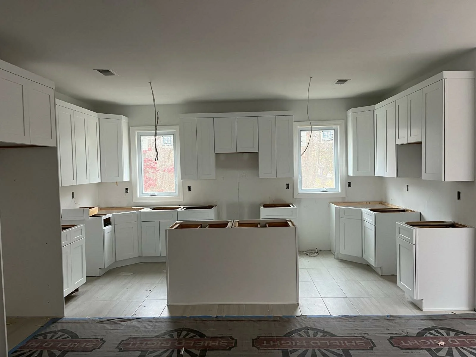 Interior of a kitchen under construction with white cabinets and two windows, no countertops installed yet.