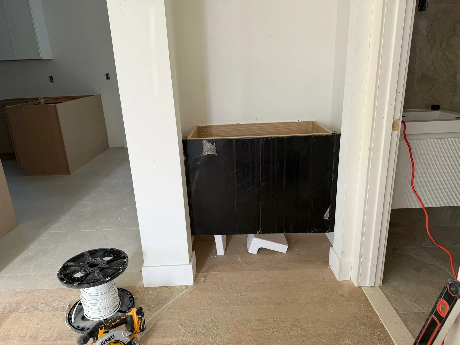 Kitchen area under renovation with a partially installed black cabinet, a red extension cord, a black and yellow power drill, a white wire spool, and construction materials on the floor.