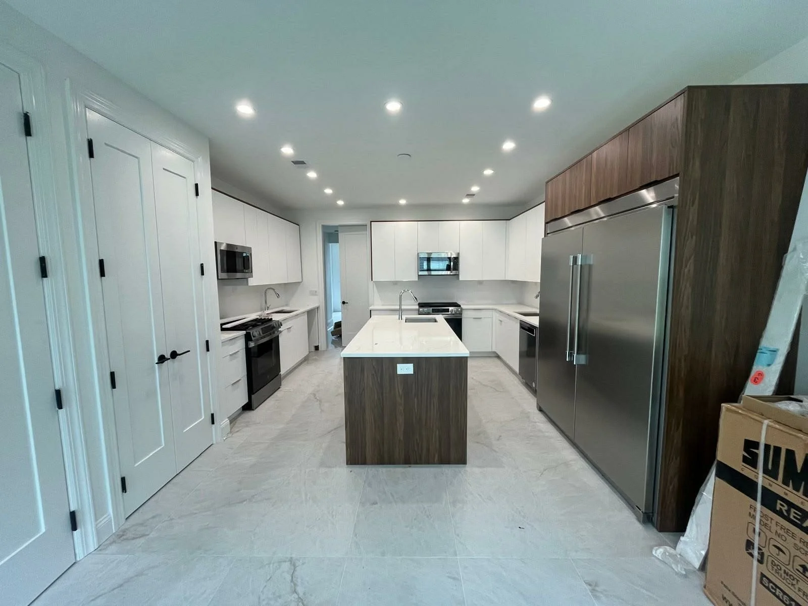 Modern kitchen with white cabinets, stainless steel appliances, a central island with a white countertop, and wood accents on the refrigerator area. There are LED recessed lights on the ceiling and a door leading to another room.