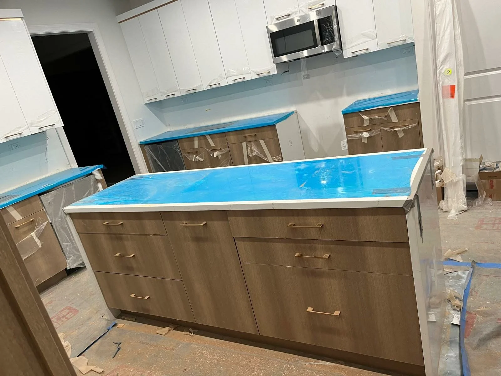 Kitchen under construction with wooden cabinets, a large central island, white upper cabinets, and blue tape covering the countertops, with construction materials and plastic coverings around.
