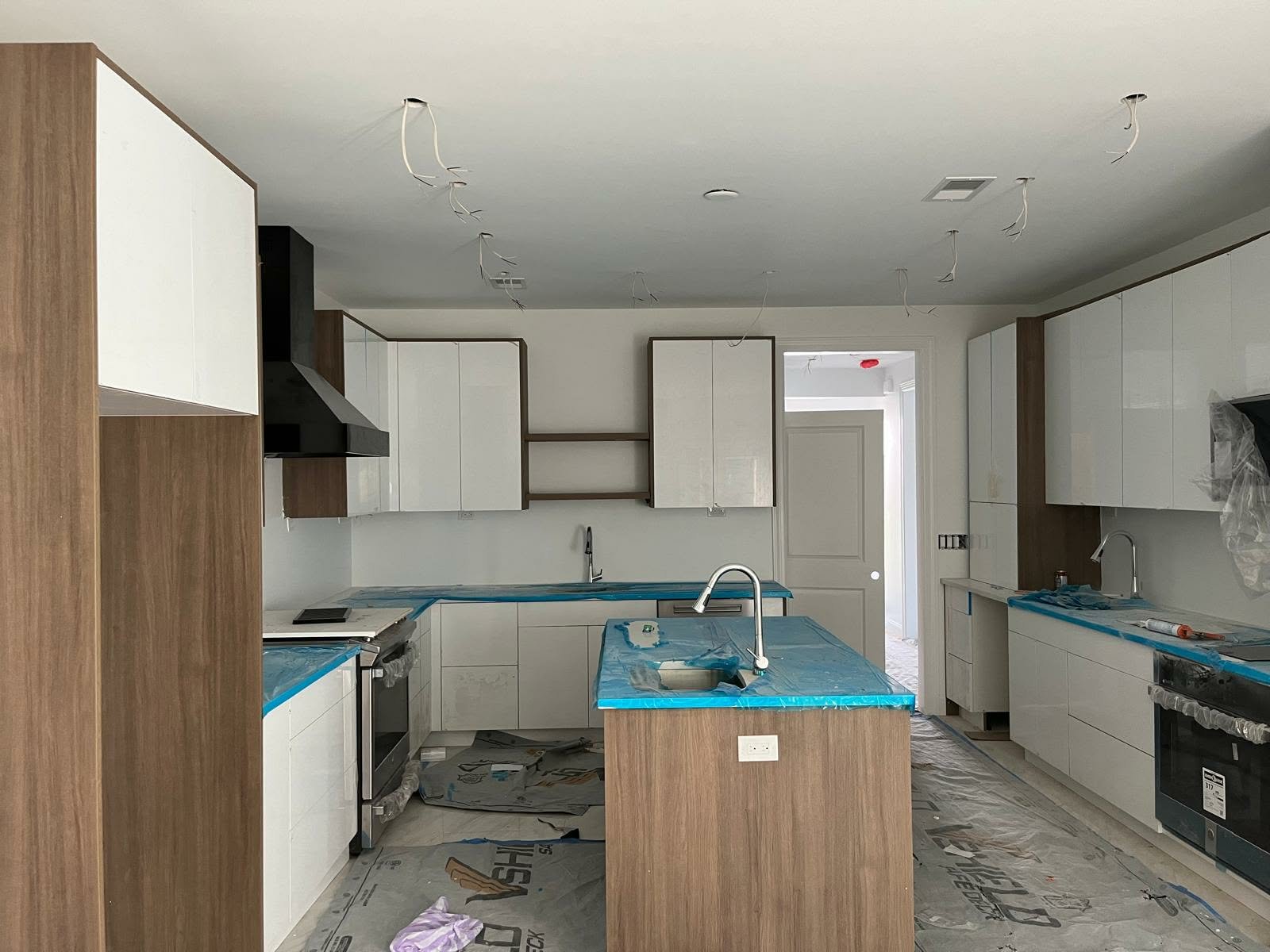 A kitchen under construction with white cabinets, black range hood, island with blue plastic covering, wooden accents, and wires hanging from the ceiling.