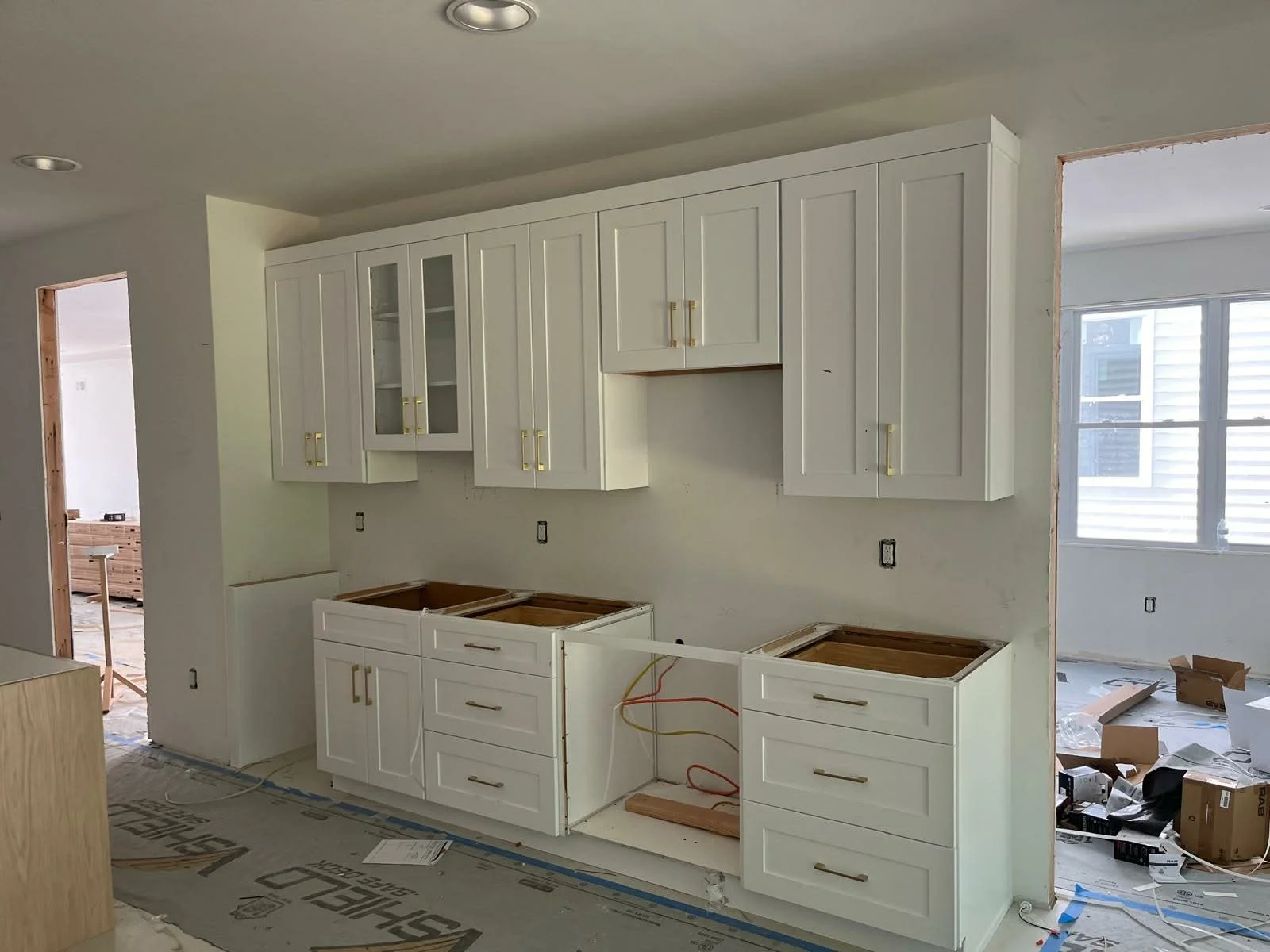Kitchen cabinetry under construction with upper cabinets mounted on the wall, lower cabinets without countertops, in a room with window and ongoing construction materials visible.
