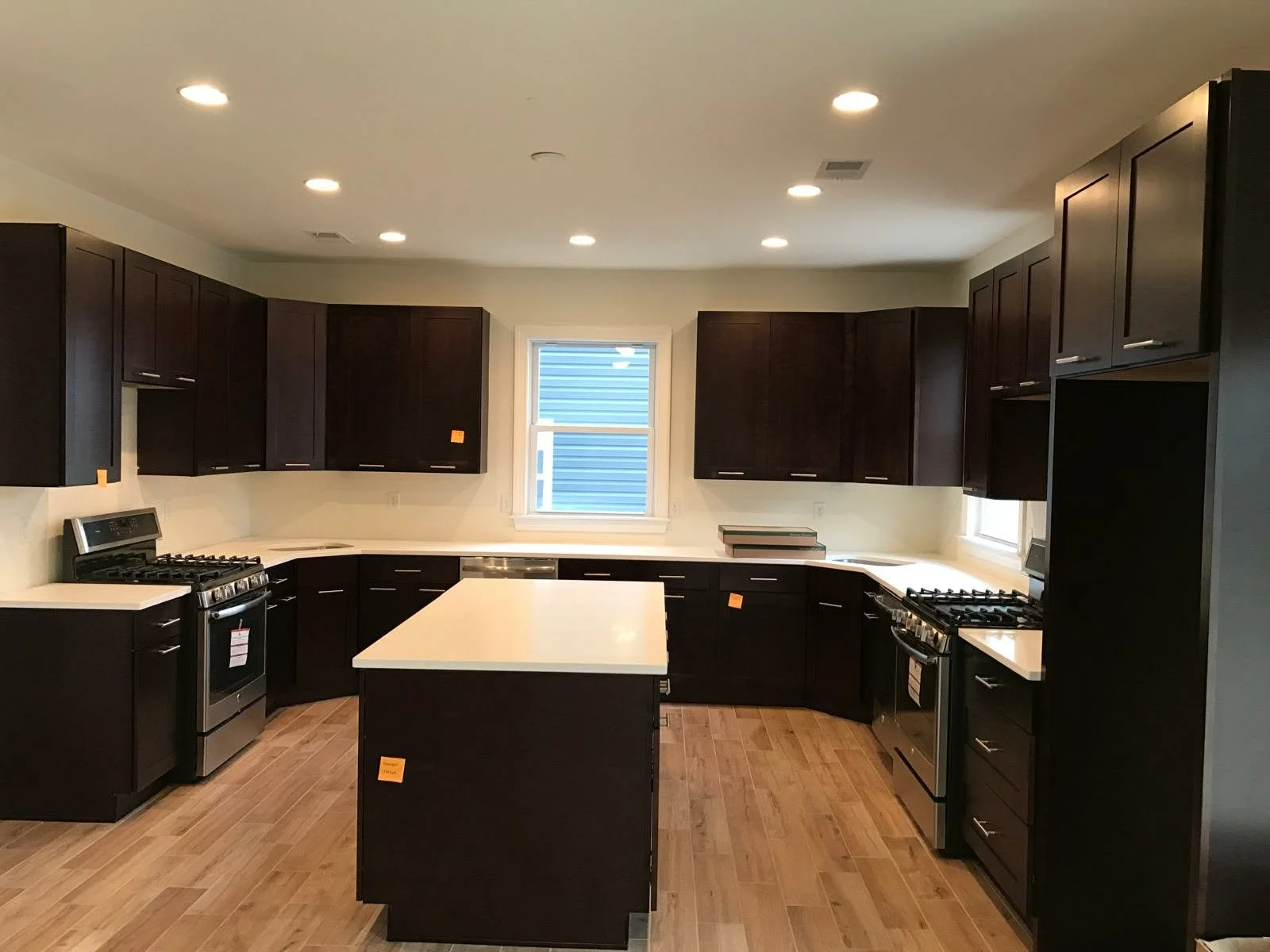 Modern kitchen with dark wood cabinets, white countertops, and stainless steel appliances, including a stove, oven, and dishwasher, with a central island, hardwood flooring, and two windows.