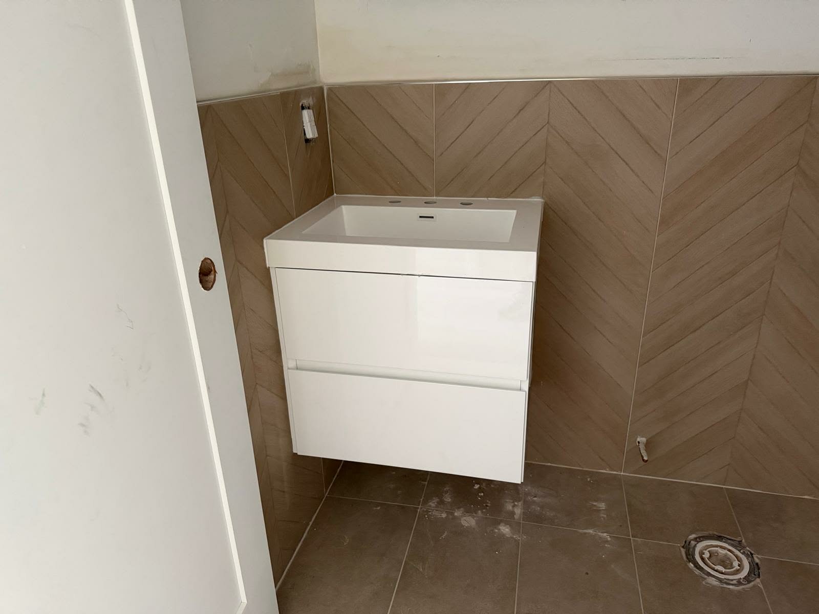 Empty bathroom with brown tiled walls and a white cabinet with a sink, no faucet installed, and a visible pipe in the floor.