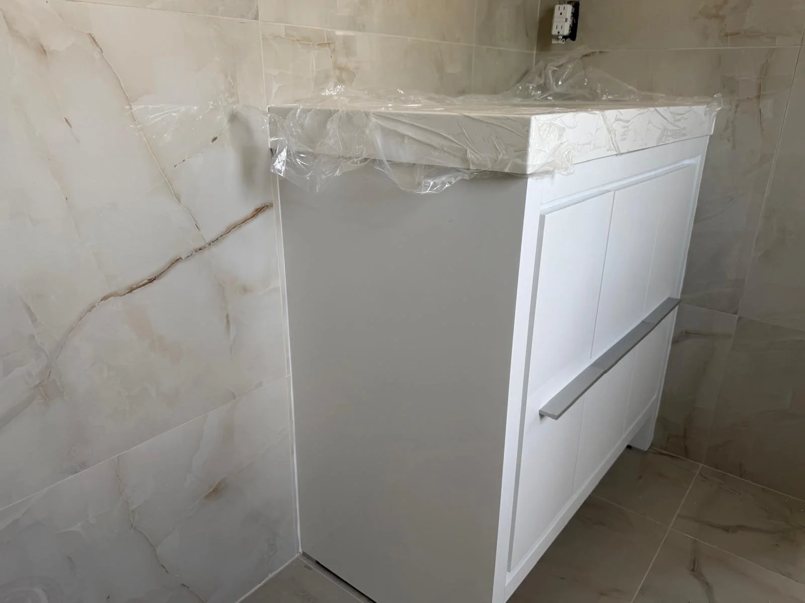 White bathroom vanity cabinet with a marble countertop and a silver handle, placed against marble-tiled walls.