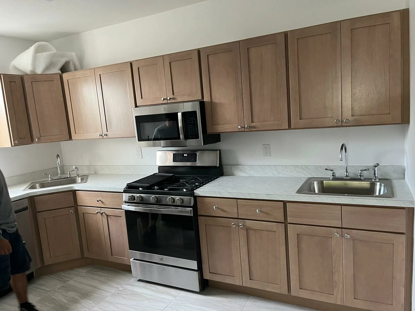 Kitchen with wooden cabinets, white countertops, stainless steel stove and microwave, two sinks, and beige tiled floor.