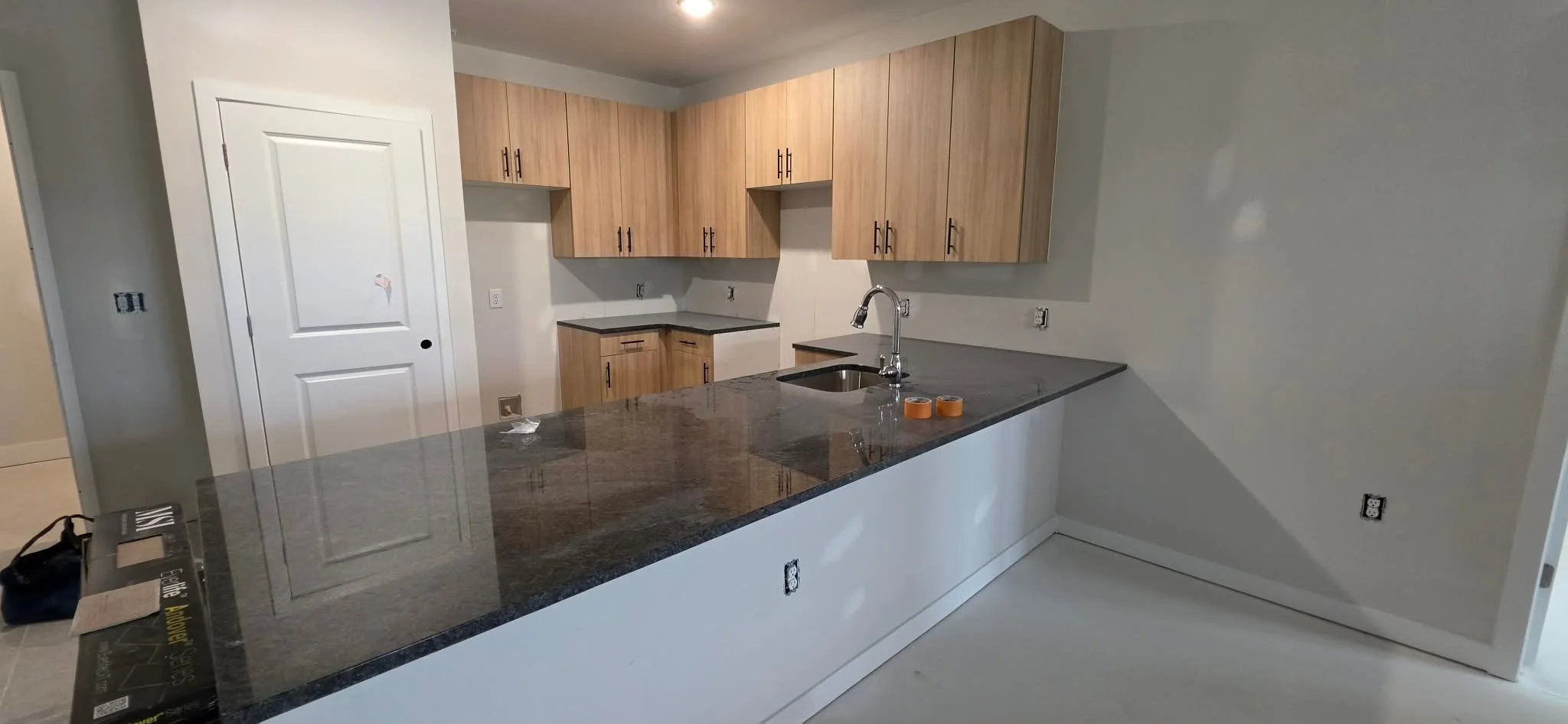Unfinished kitchen with light wood cabinets, black countertops, a stainless steel sink, and various electrical outlets and wires waiting for appliances and fixtures.