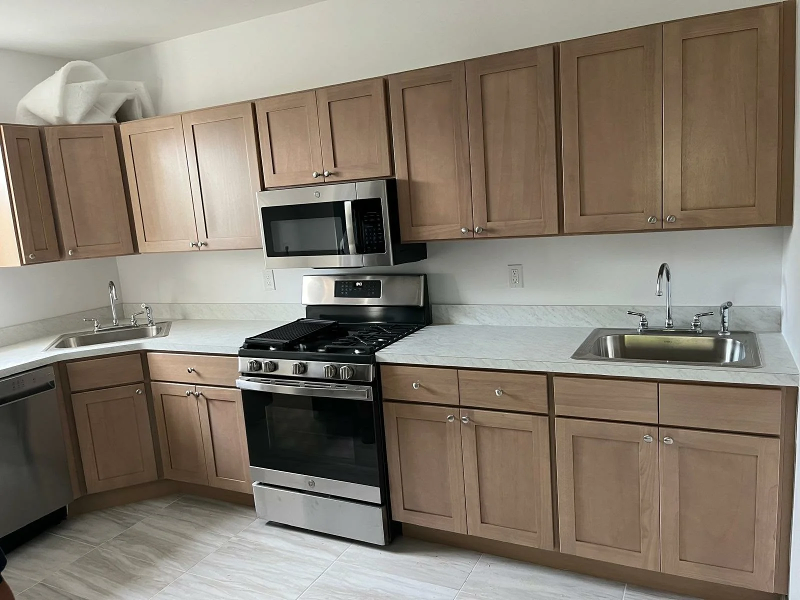 Kitchen with wooden cabinets, stainless steel microwave and oven, black stovetop, double sinks, light-colored countertops, and tile flooring.
