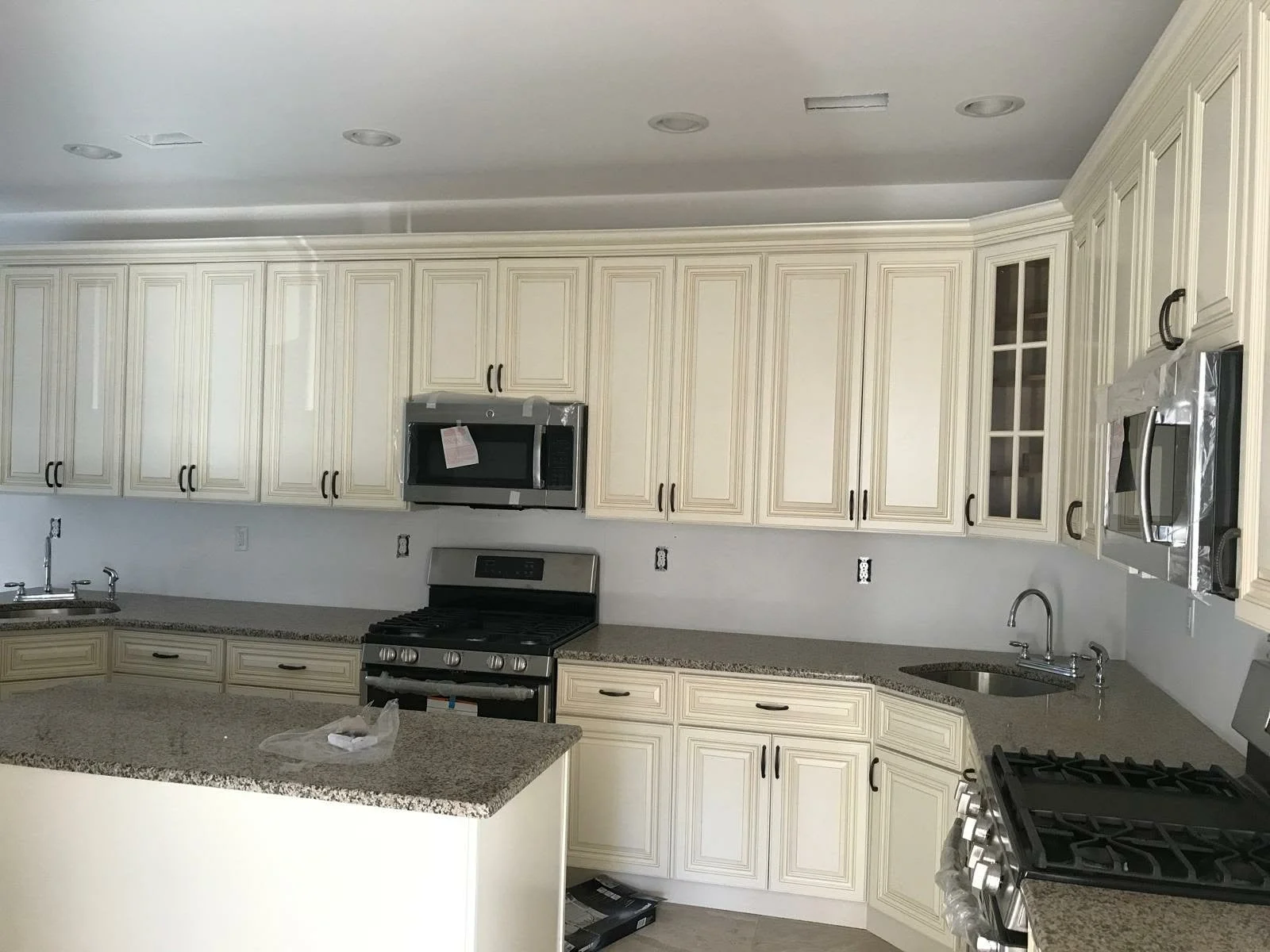 Kitchen with cream-colored cabinets, granite countertops, stainless steel appliances including a microwave, oven, and refrigerator, and a double sink under a window.