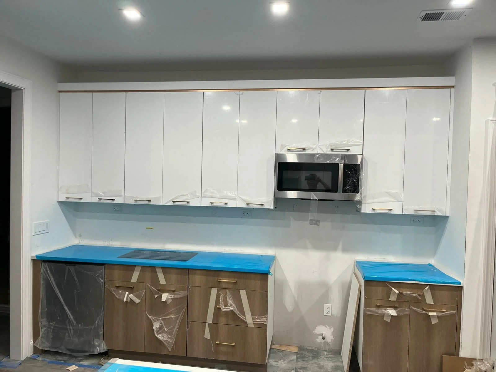 Kitchen with white upper cabinets, blue and brown lower cabinets, and a microwave still wrapped in plastic, indicating ongoing installation or renovation.