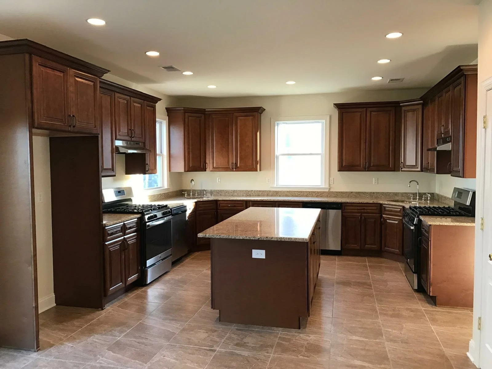 A spacious modern kitchen with dark wood cabinets, granite countertops, a kitchen island, and stainless steel appliances, including a stove, dishwasher, and oven. There are two windows allowing natural light to enter.