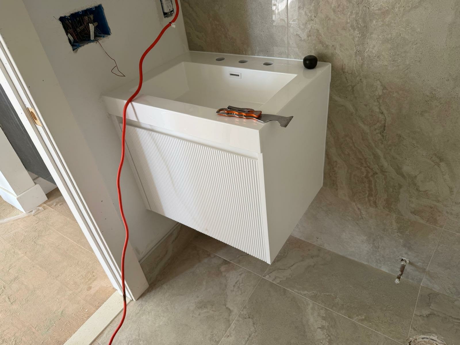 Bathroom under construction with a partially installed white vanity, a black round object on top, tools including a putty knife, and exposed electrical wiring on the wall.