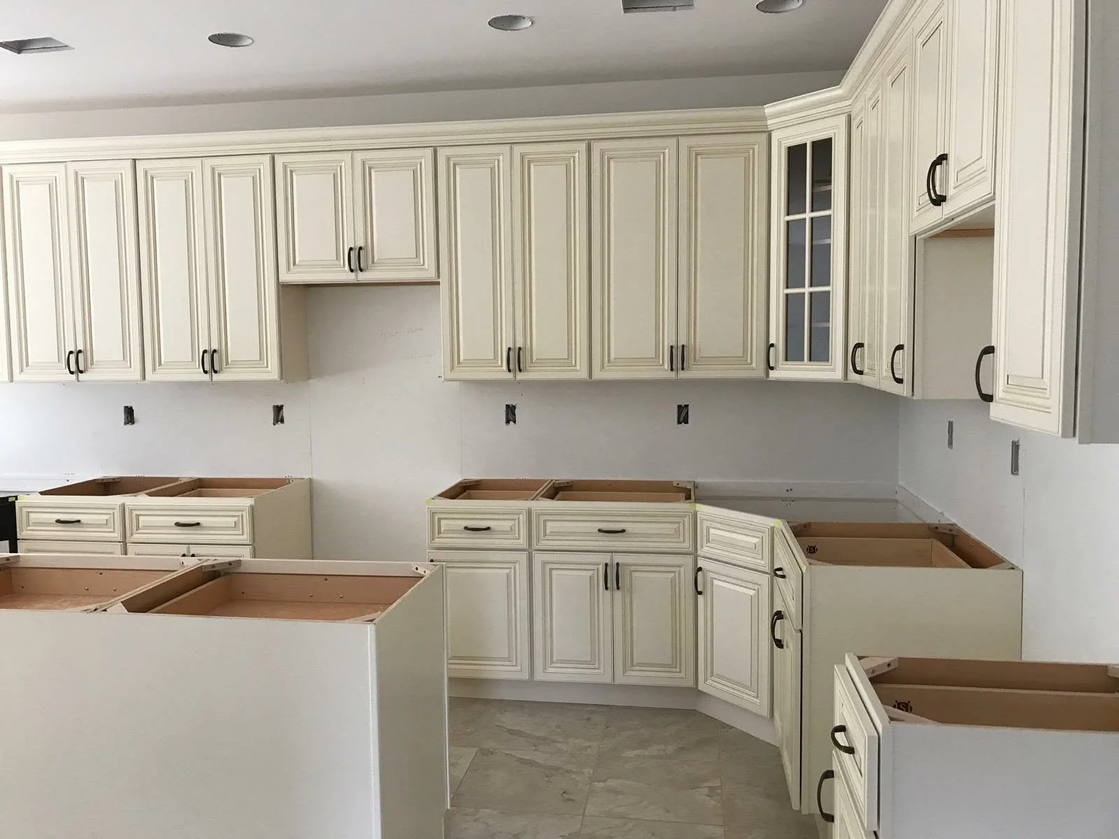 Kitchen cabinets under installation, with some lower cabinets missing countertops, in a kitchen with white cabinetry and a tiled floor.