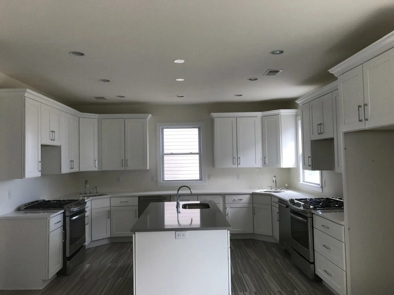 Modern kitchen with white cabinets, gray countertops, stainless steel appliances, and hardwood flooring. There are two windows bringing in natural light.