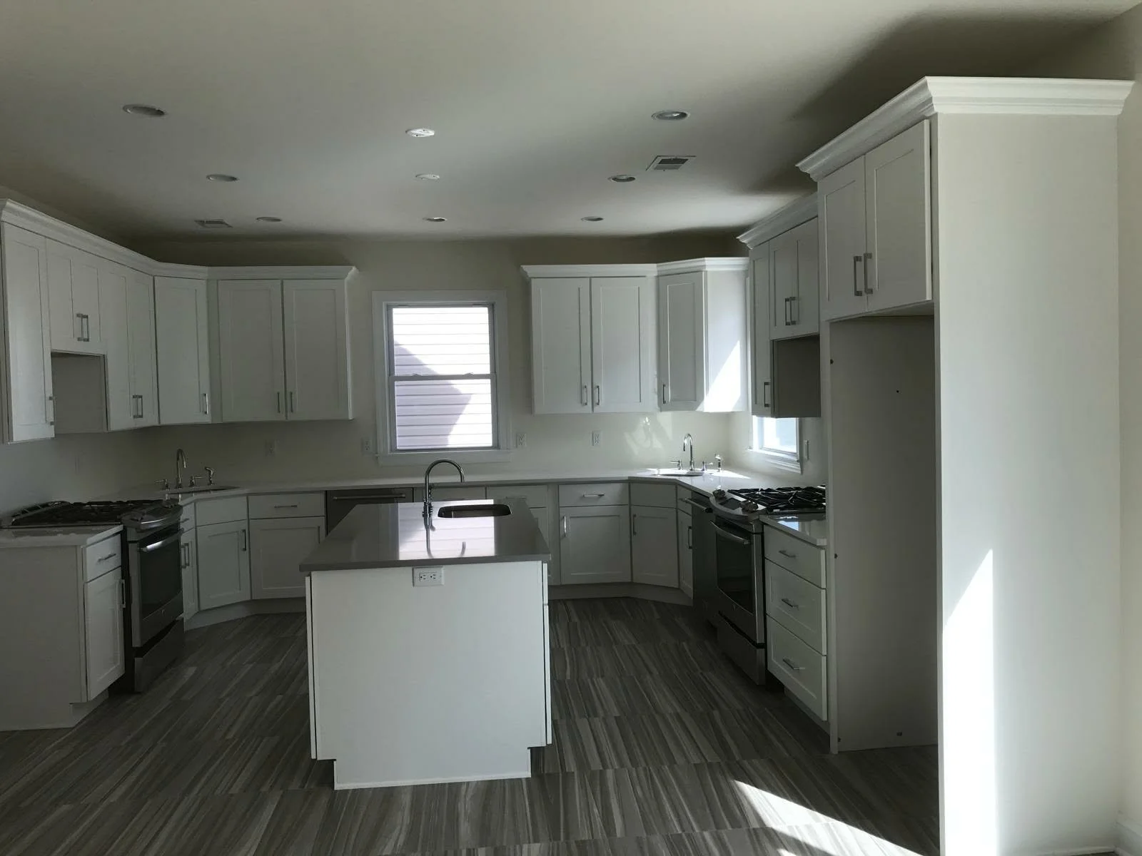 Empty kitchen with white cabinets, gray countertops, stainless steel appliances, and a central island with a sink, illuminated by natural light from two windows.