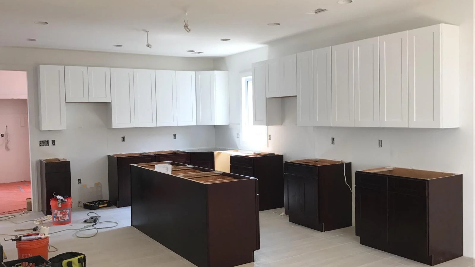 Kitchen under construction with white upper cabinets, dark lower cabinets, and no countertops installed.