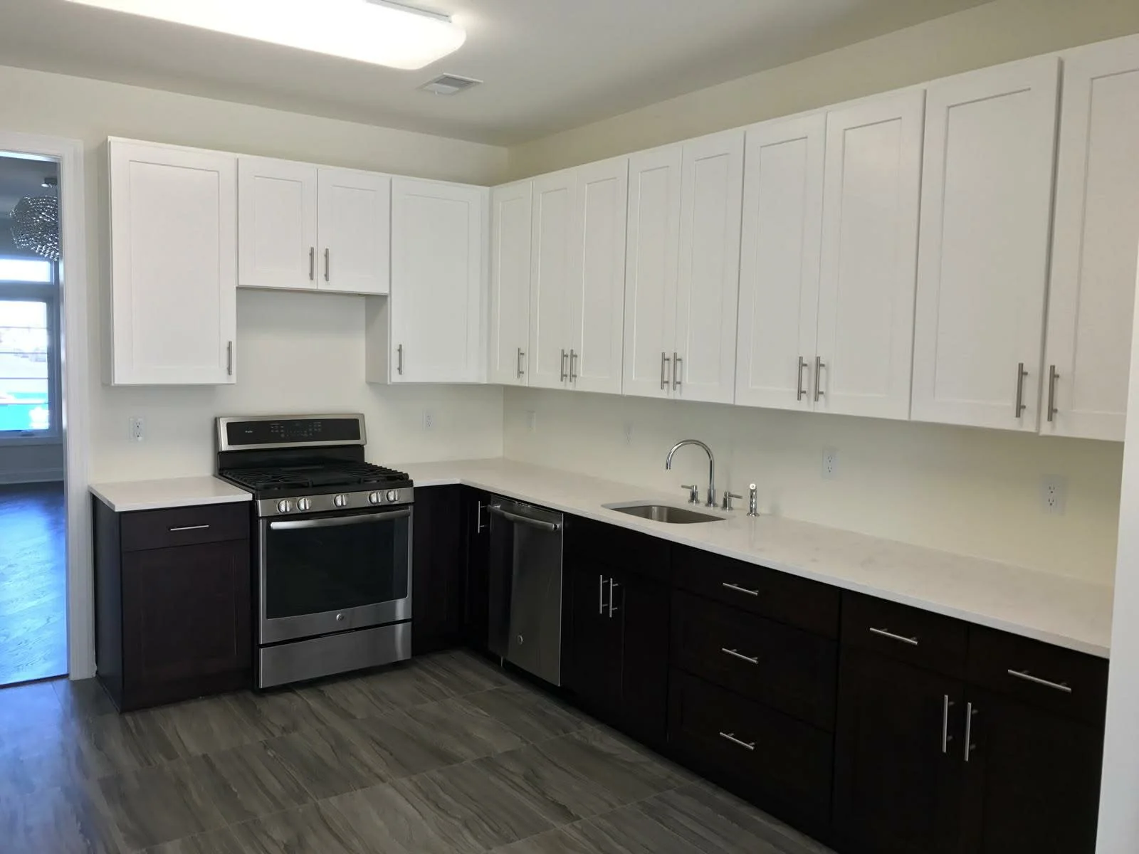Modern kitchen with white upper cabinets, dark lower cabinets, stainless steel appliances, including stove, dishwasher, and fridge, with a sink under the window, and wood-look tile flooring.