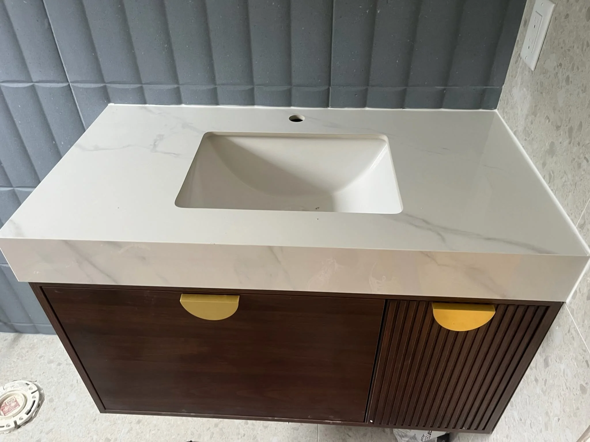 A bathroom vanity with a white marble countertop, an integrated sink, and a dark wood cabinet with rounded yellow handles. Behind it, there's a wall covered in gray textured tiles, and a power outlet is visible on the right side. The floor is light-colored with a tiled pattern.